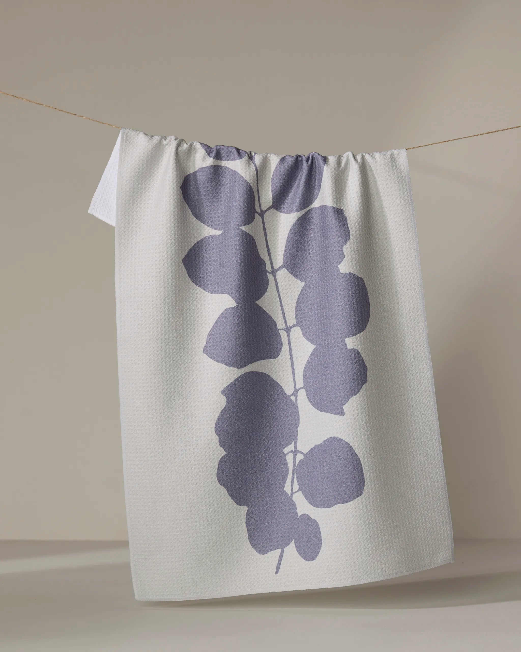 The PrintedPlant Carob tea towel, featuring a bold, soft purple botanical leaf print, hangs on a line against a neutral background.