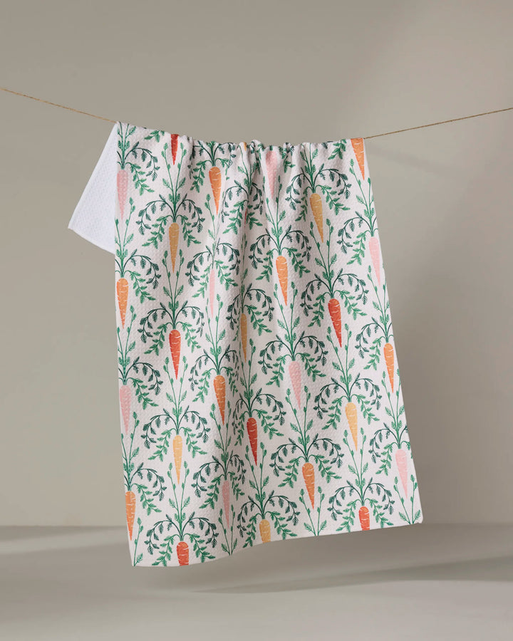 The "Carrot Damask" dish towel by Sarah Daugherty, featuring an illustrated orange carrot and green leaf pattern, hangs on a clothesline against a neutral background.