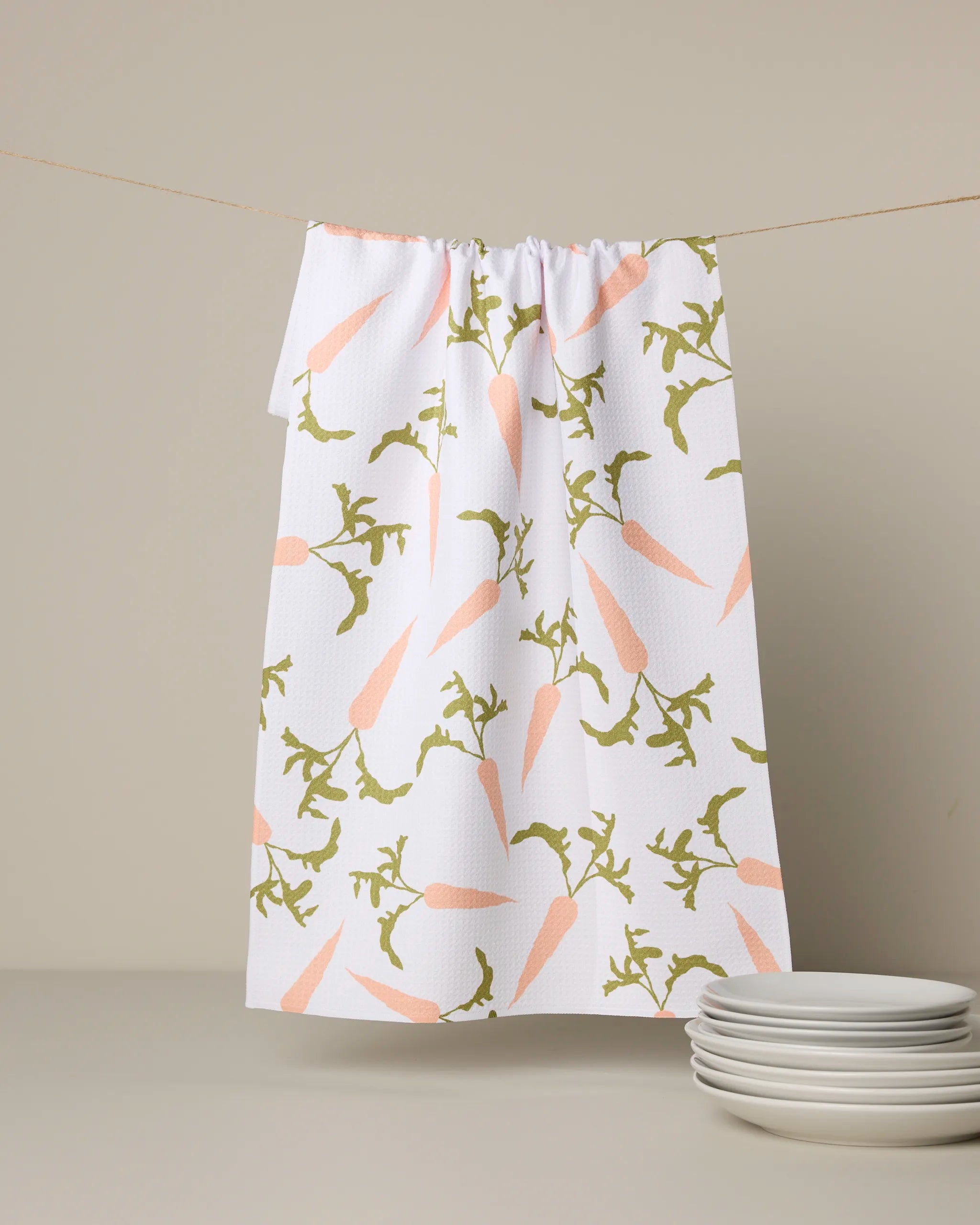 The Geometry Carrot Toss recycled kitchen towel, featuring orange carrots and green leaves, hangs on a clothesline above stacked white plates against a neutral background.