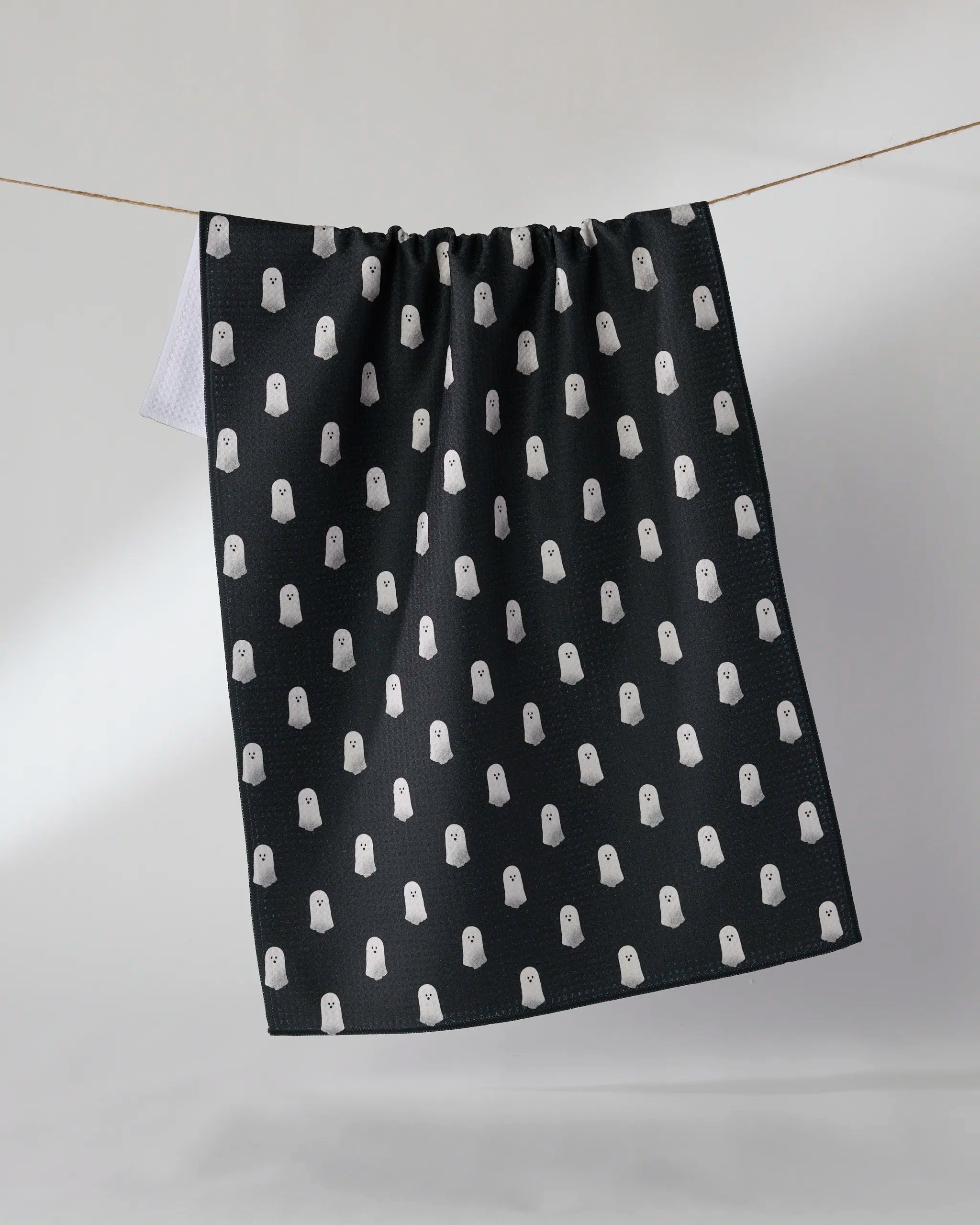 The Casper Boo tea towel by Geometry hangs on a clothesline, displaying its black fabric and playful white cartoon ghost pattern. Made from recycled materials, this ultra-absorbent towel blends fun design with eco-friendly practicality.