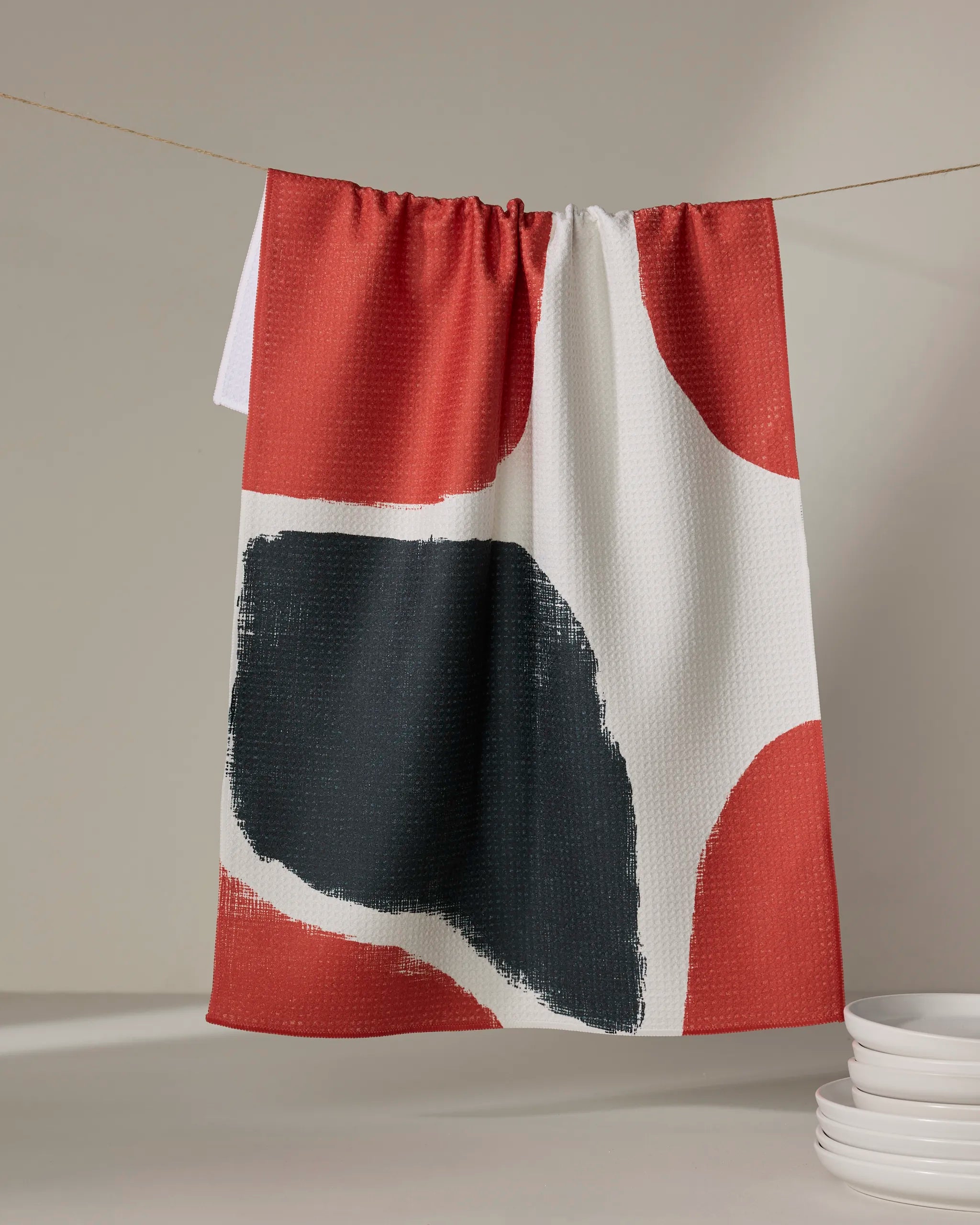 The Geometry Cast tea towel, featuring a quick-drying abstract design in red, black, and white, hangs on a string with a stack of white plates nearby on a light surface.