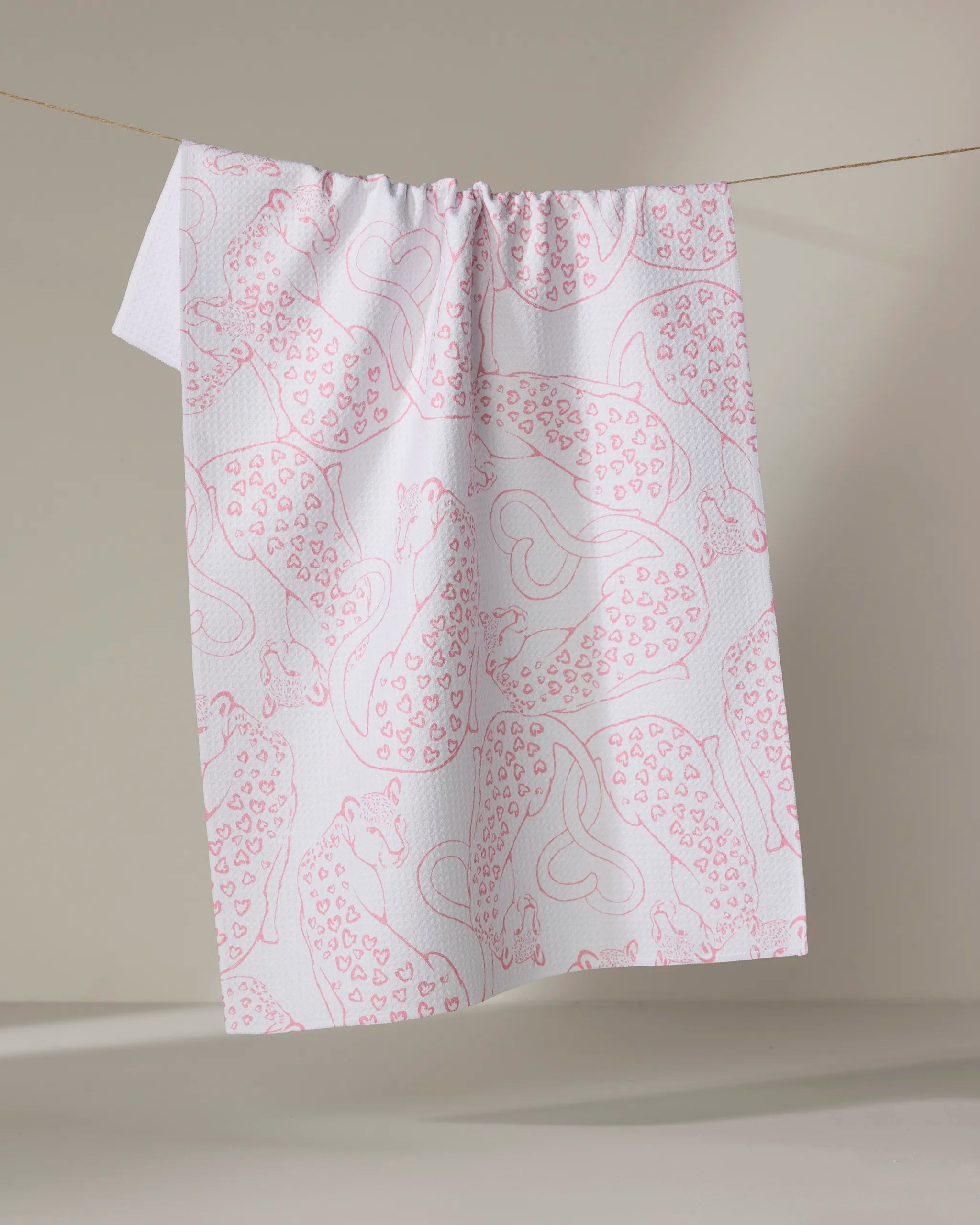 The Cat Lover tea towel by Hufton Studio, featuring pink outlined leopards and hearts on white recycled fabric, hangs on a string against a neutral background.
