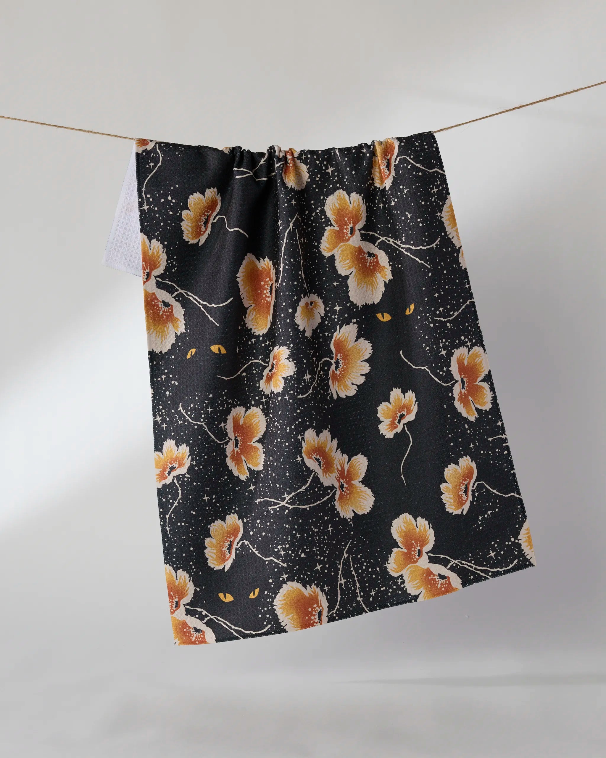 The Cats Eye tea towel by Geometry features an orange and white floral pattern with yellow cat eyes on black, made from recycled materials, and is shown hanging against a light background.