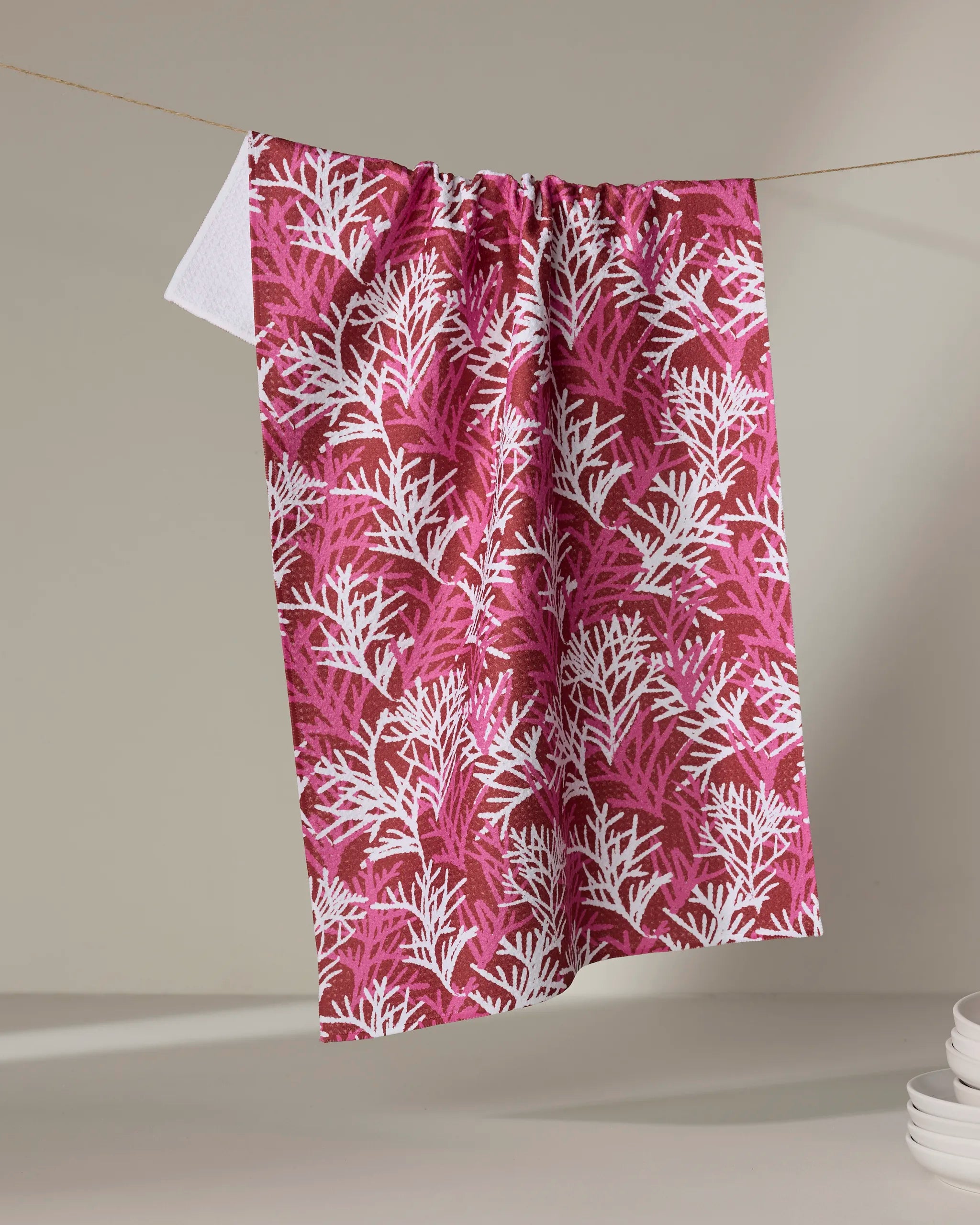 The Kayla Ann Cedar Pine kitchen tea towel, crafted from recycled materials with a coral reef pattern in red and white, hangs from a clothesline beside a stack of white plates.