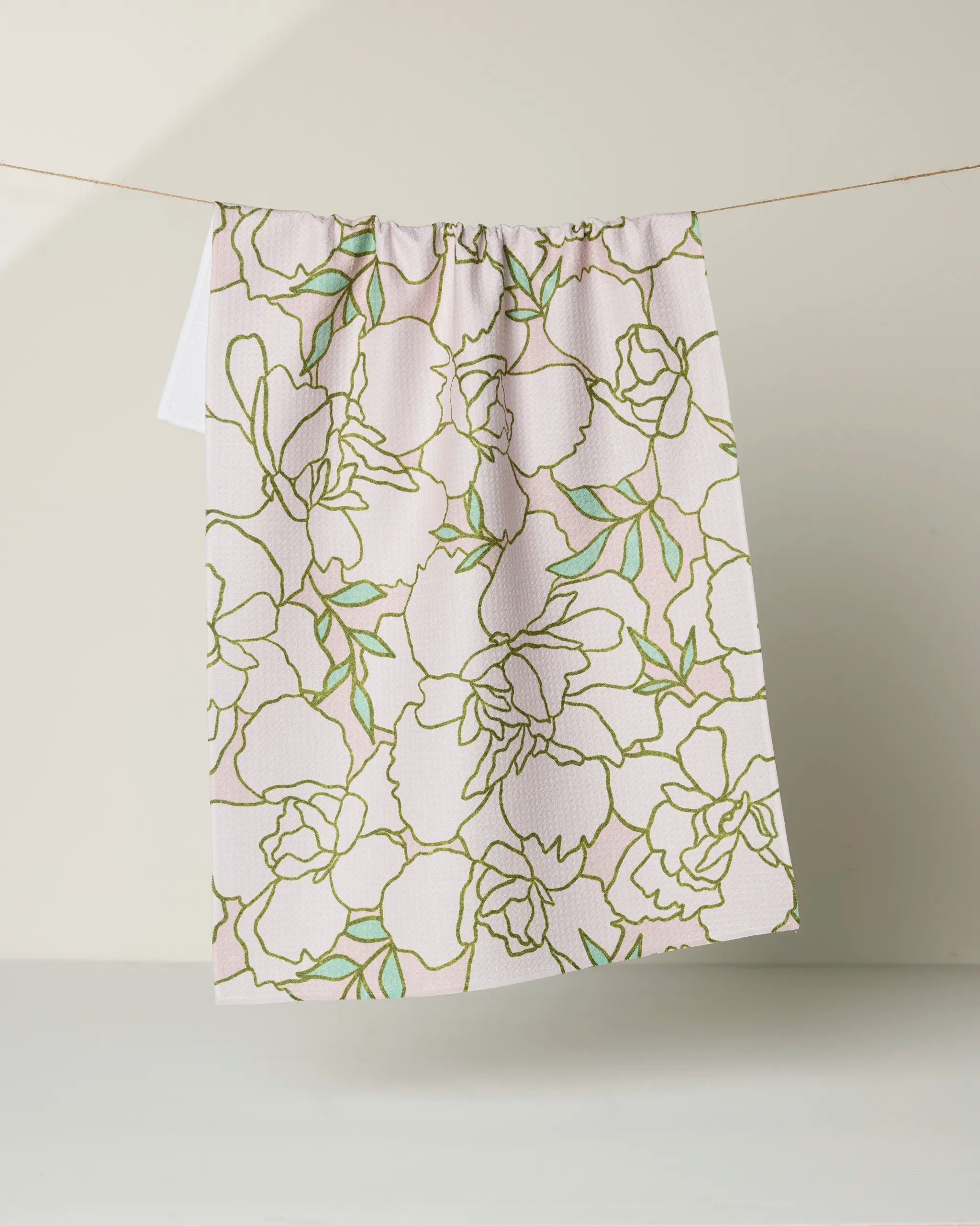 The Celecia kitchen towel by Amy MacCready, featuring a green and yellow floral line pattern on white, hangs from a string against a plain, light background.