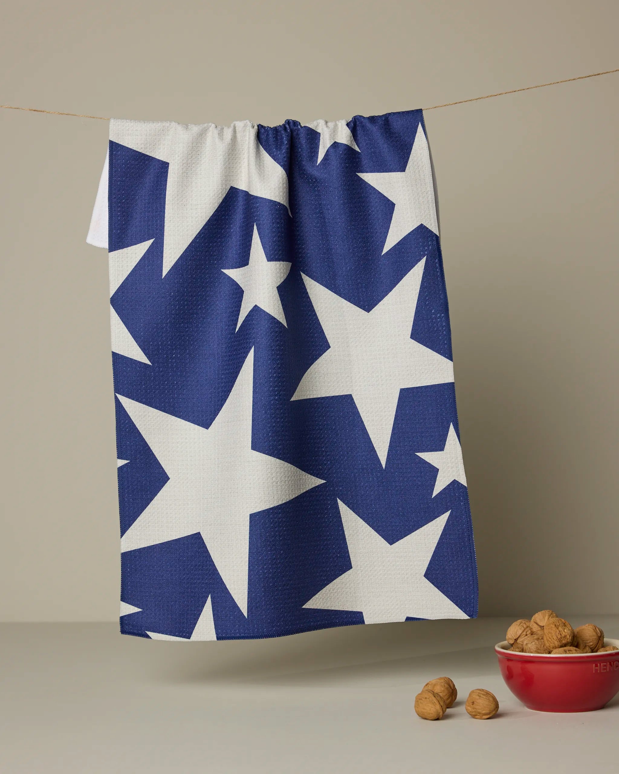 The Celeste kitchen towel by Geometry, featuring large white stars on blue recycled fabric, hangs on a line above a red bowl of walnuts and some loose walnuts on a light surface with a simple background.