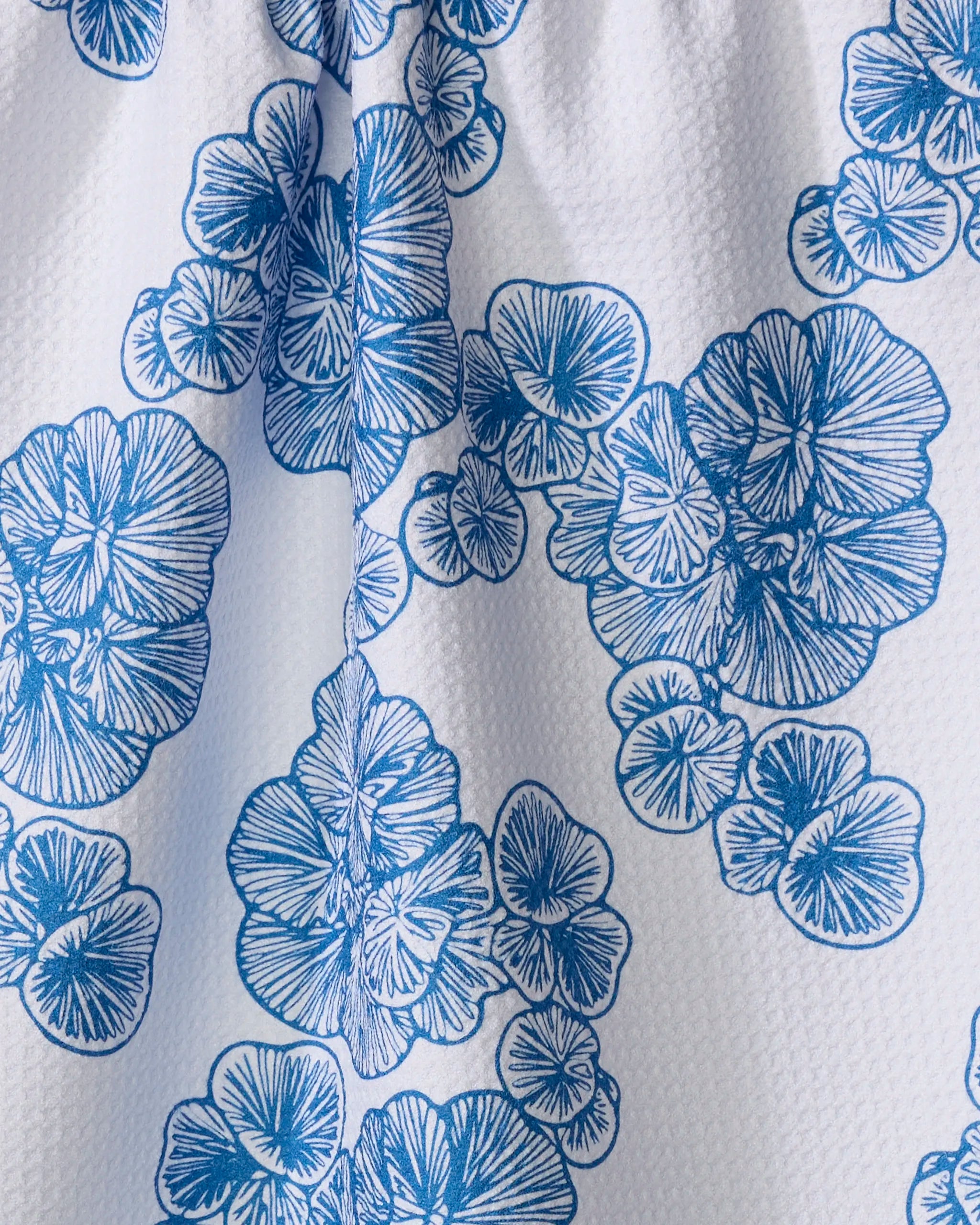 The Celia tea towel by Second Nature Studio is a white, textured kitchen towel with a blue floral pattern of stylized overlapping flowers, detailed line work, and a vertical fold down the center.
