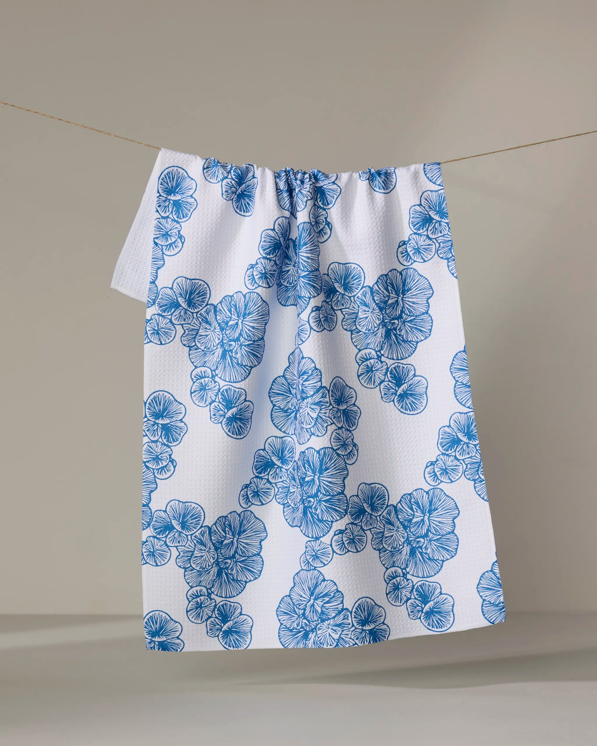 The Celia kitchen towel by Second Nature Studio hangs on a line, showcasing a blue clustered floral pattern on a white, textured surface. Made from recycled materials, it’s set against a softly lit, neutral background.