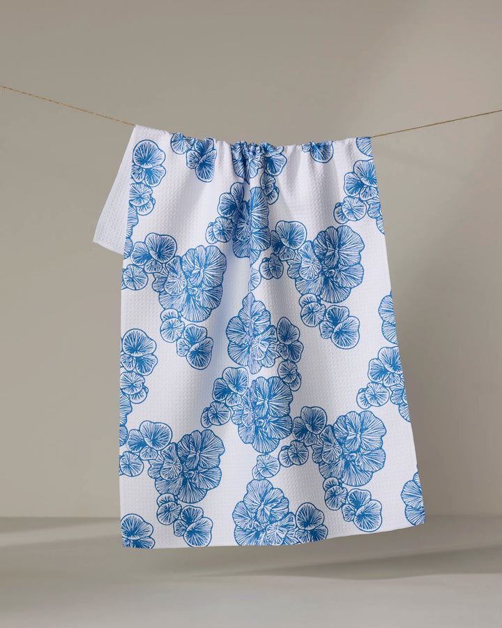 The Celia kitchen towel by Second Nature Studio hangs on a line, showcasing a blue clustered floral pattern on a white, textured surface. Made from recycled materials, it’s set against a softly lit, neutral background.