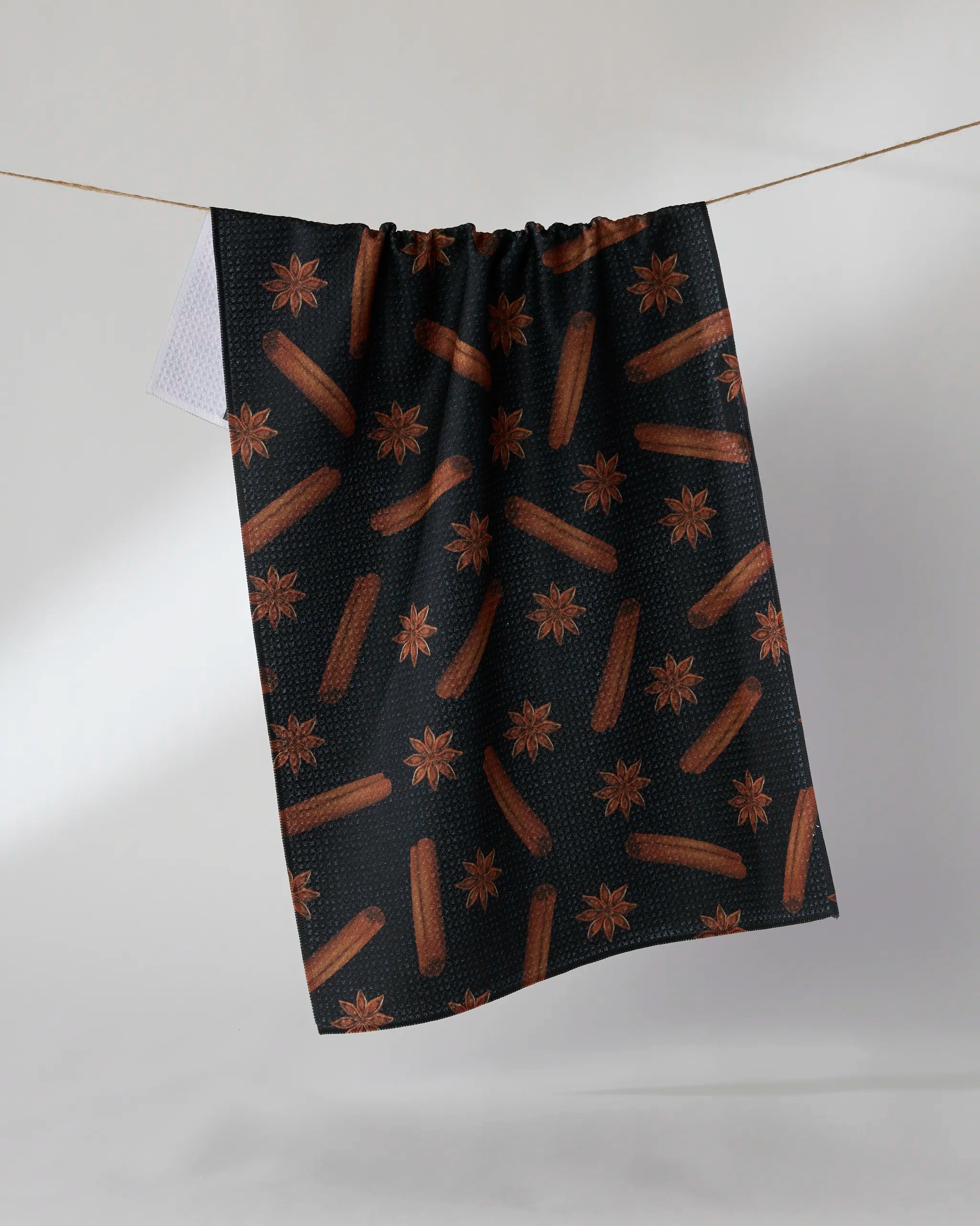 The Chai Noir tea towel by Geometry, featuring brown star anise and cinnamon sticks on a dark background, hangs on a clothesline against a plain light backdrop.