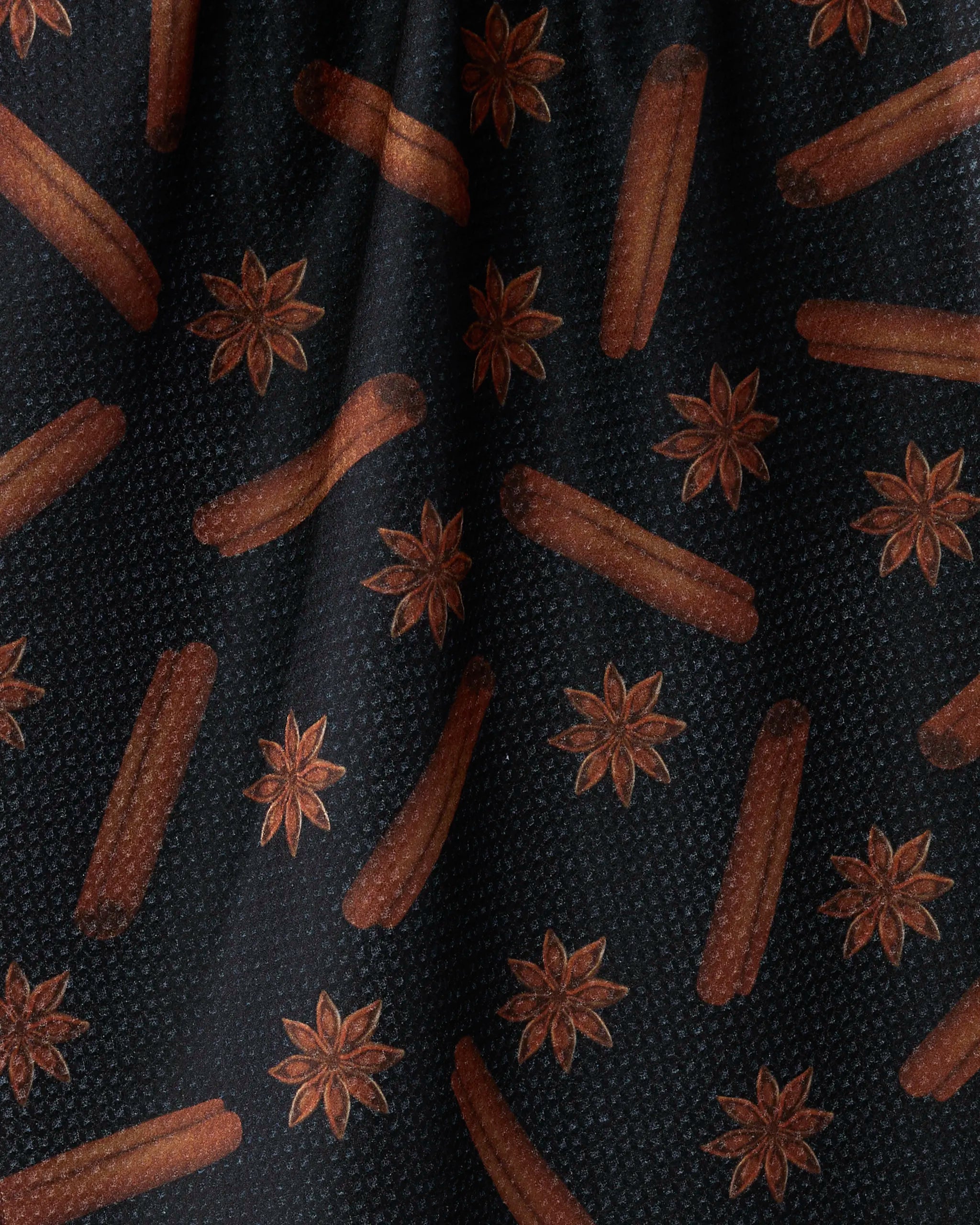 The Chai Noir tea towel by Geometry features a dark textured background with repeating brown churros and star anise illustrations, crafted from recycled materials.