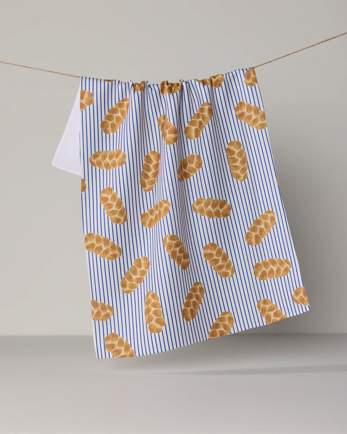 The “Challah Days” tea towel by Geometry features blue and white stripes adorned with a playful pattern of brown braided challah loaves, hanging on a clothesline against a neutral backdrop. This eco-friendly kitchen towel blends charming design with practical use, making it perfect for any home baker.