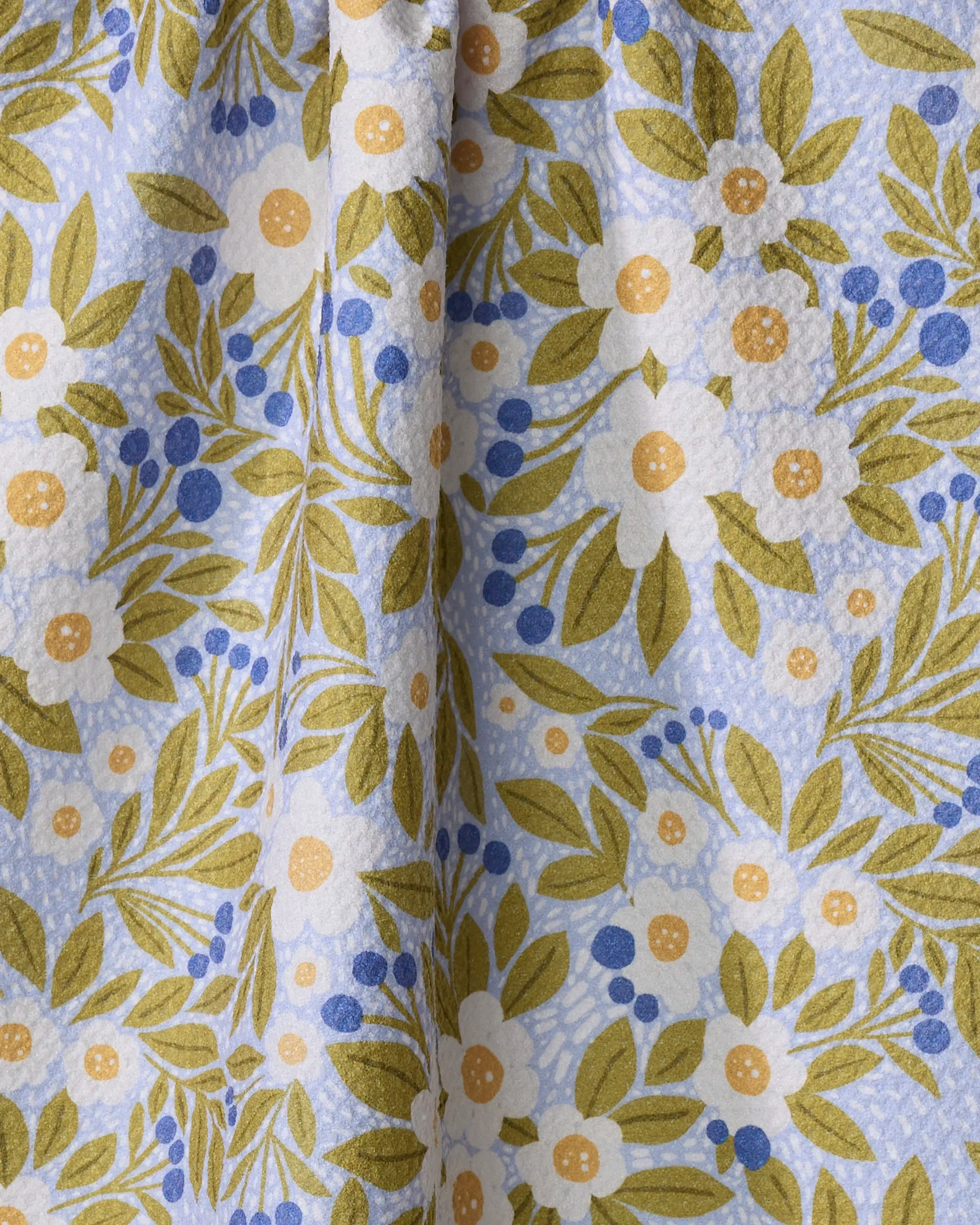 A close-up of Lisa Nelson’s Chamomile And Blue tea towel, showing green leaves, white and yellow flowers, and tiny blue berries on a light blue background. The ultra-absorbent fabric is softly gathered at the center.
