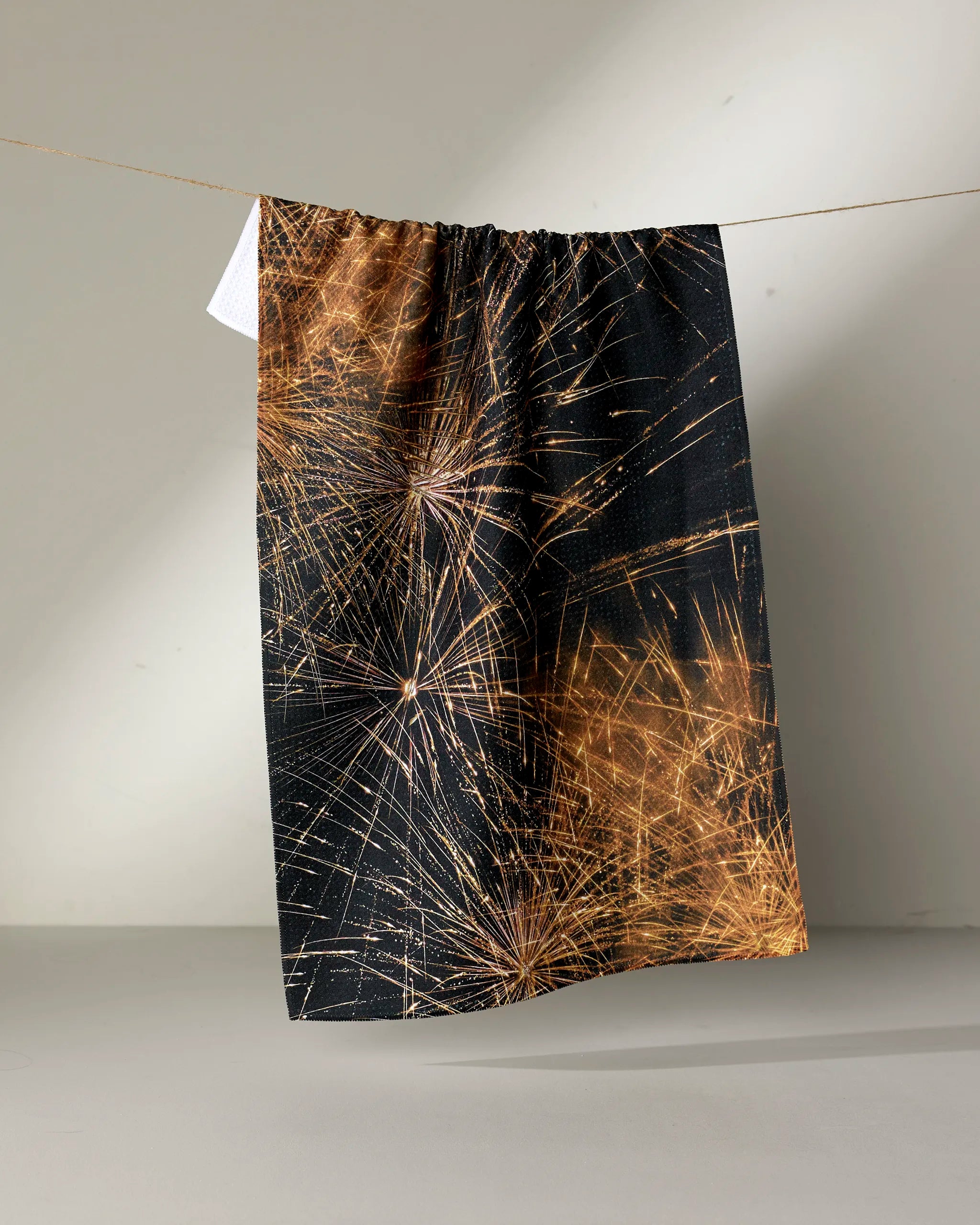The Geometry "Champagne Sparkler" tea towel, featuring a black background with gold fireworks, hangs on a clothesline against a plain, light-colored wall.