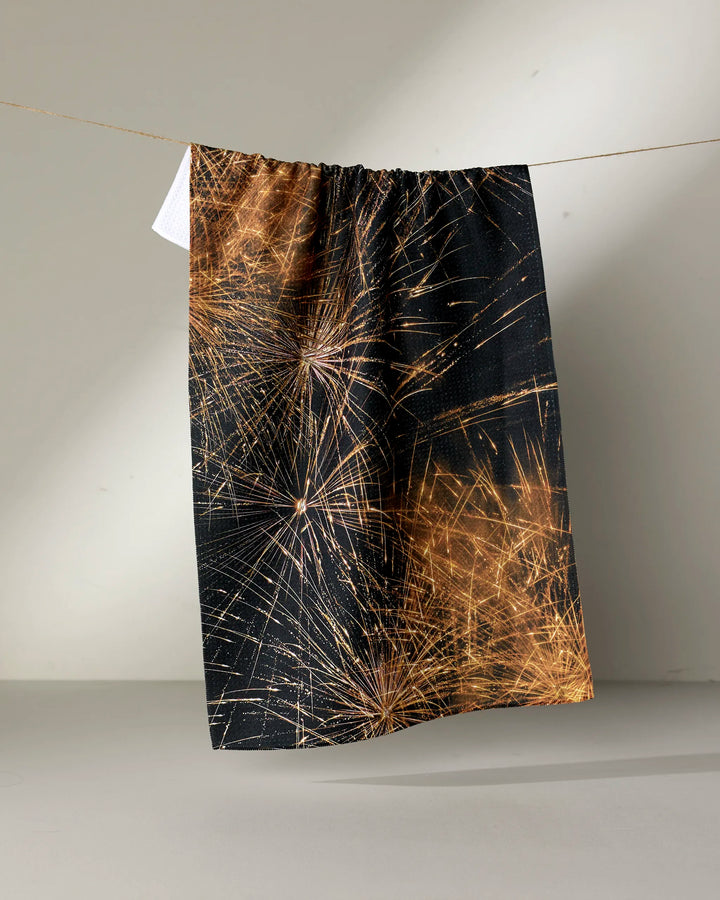 The Geometry "Champagne Sparkler" tea towel, featuring a black background with gold fireworks, hangs on a clothesline against a plain, light-colored wall.