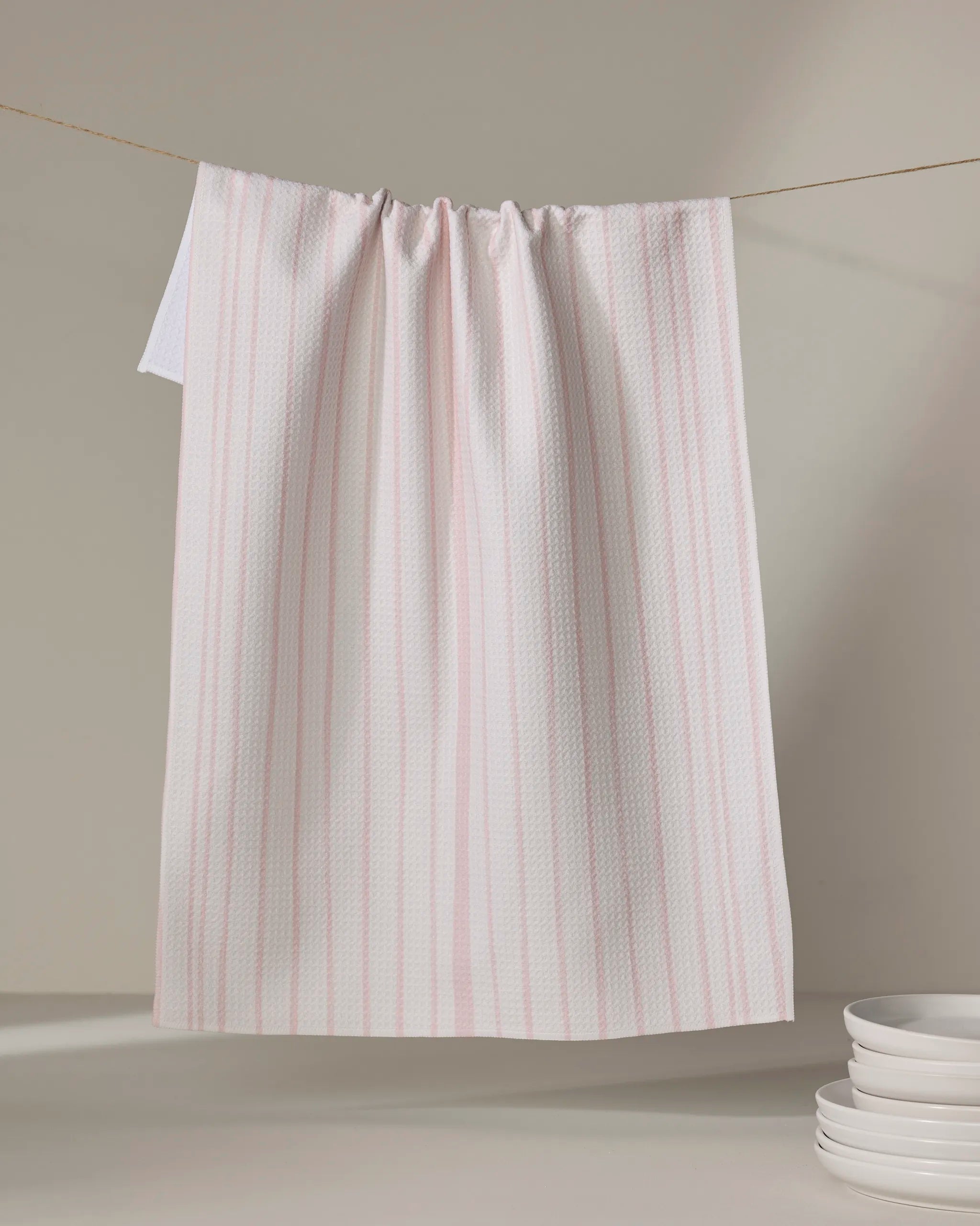 The Becki Owens Charlotte Stripe tea towel, featuring subtle pink vertical stripes on white, hangs on a string against a neutral background, with white plates stacked in the lower right corner.