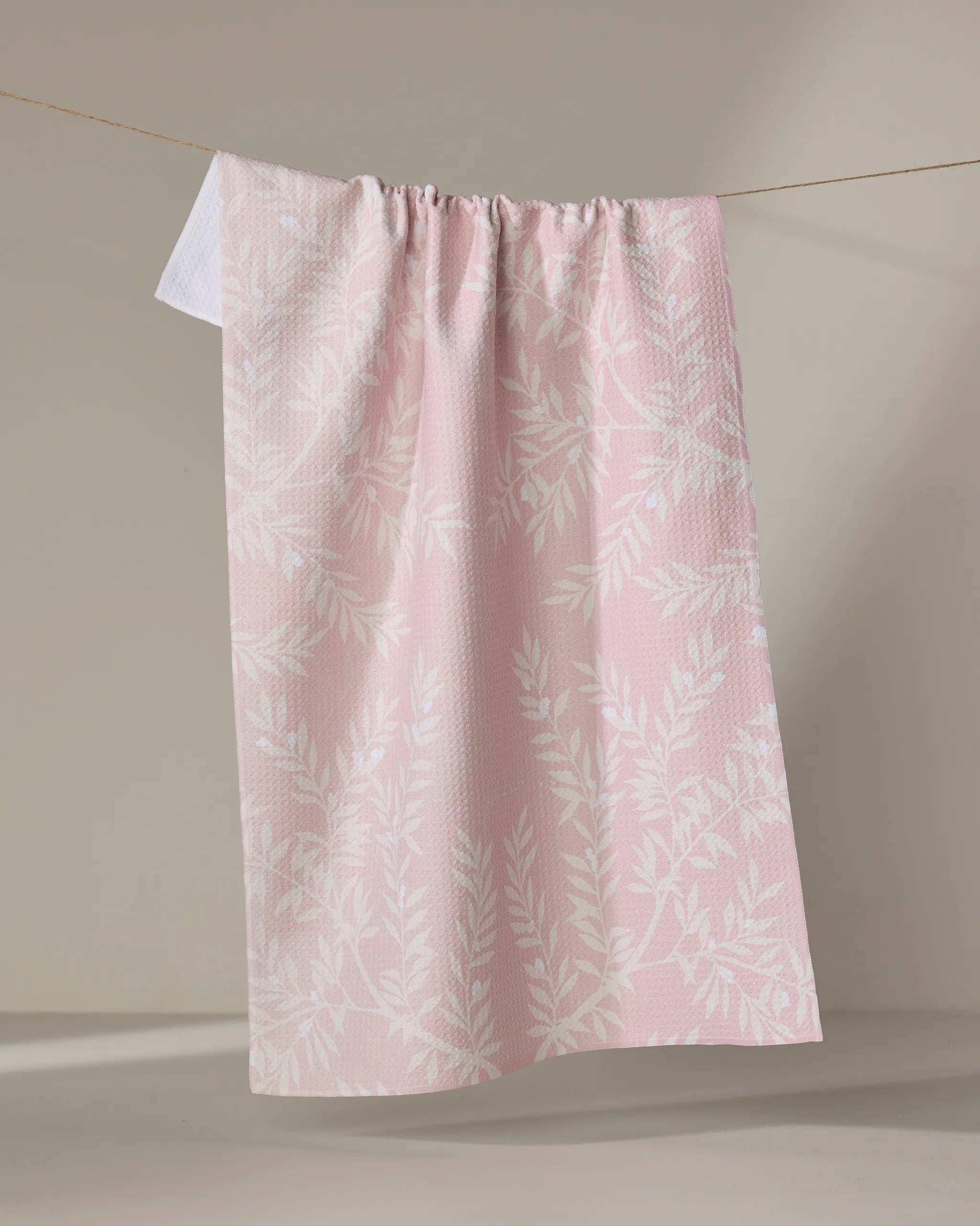 The Becki Owens Charlotte kitchen towel, in light pink with a subtle white leafy pattern, hangs on a clothesline against a neutral background.