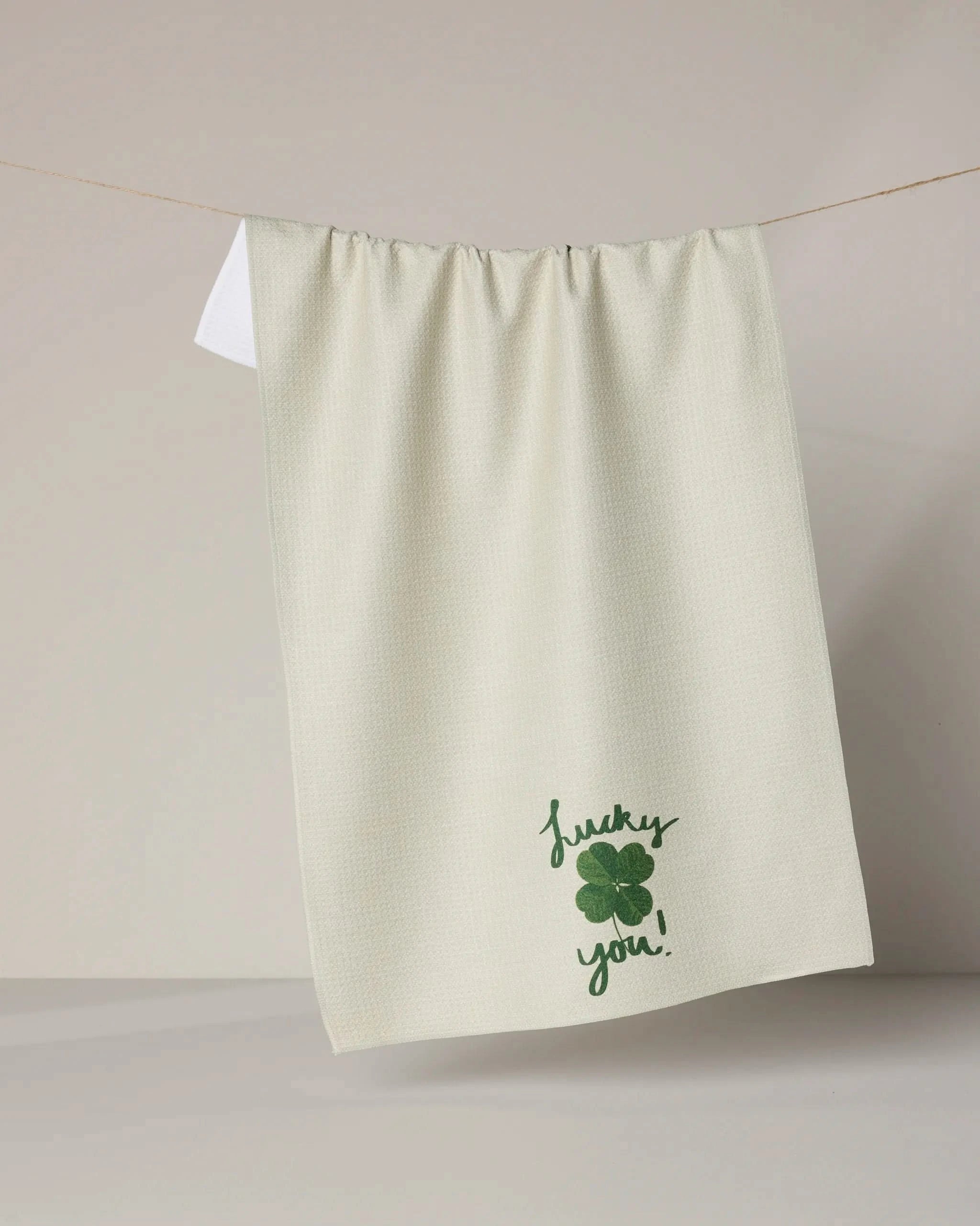 A cream-colored recycled kitchen towel from Geometry’s Charmed Up collection hangs on a line, featuring a green four-leaf clover and “lucky you!” embroidered near the bottom.