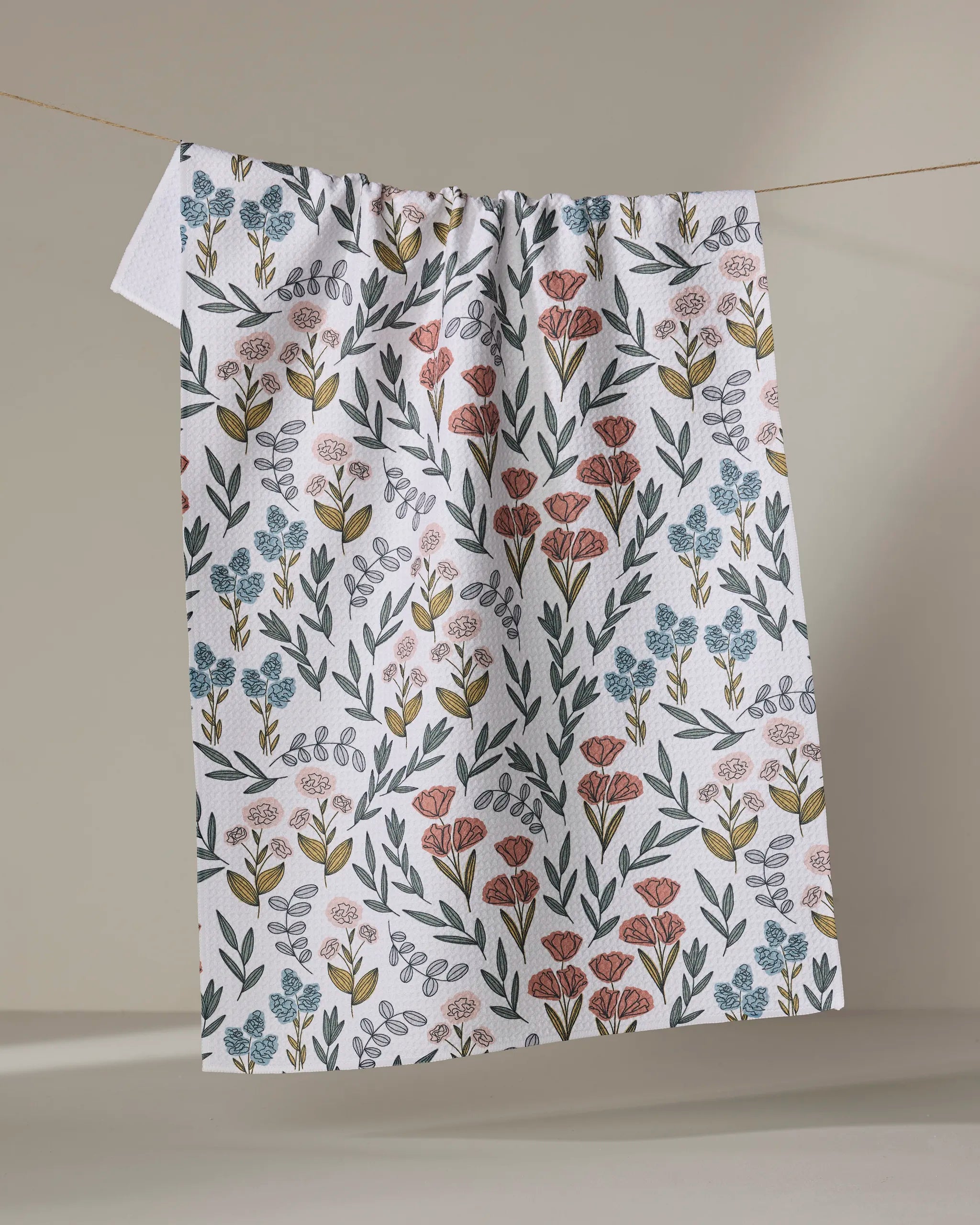 The Charolette Multi tea towel by Charlie Rowan Designs, made from ultra-absorbent recycled materials, features a vibrant floral pattern with red, blue, and yellow flowers and green leaves and hangs on a clothesline against a neutral background.