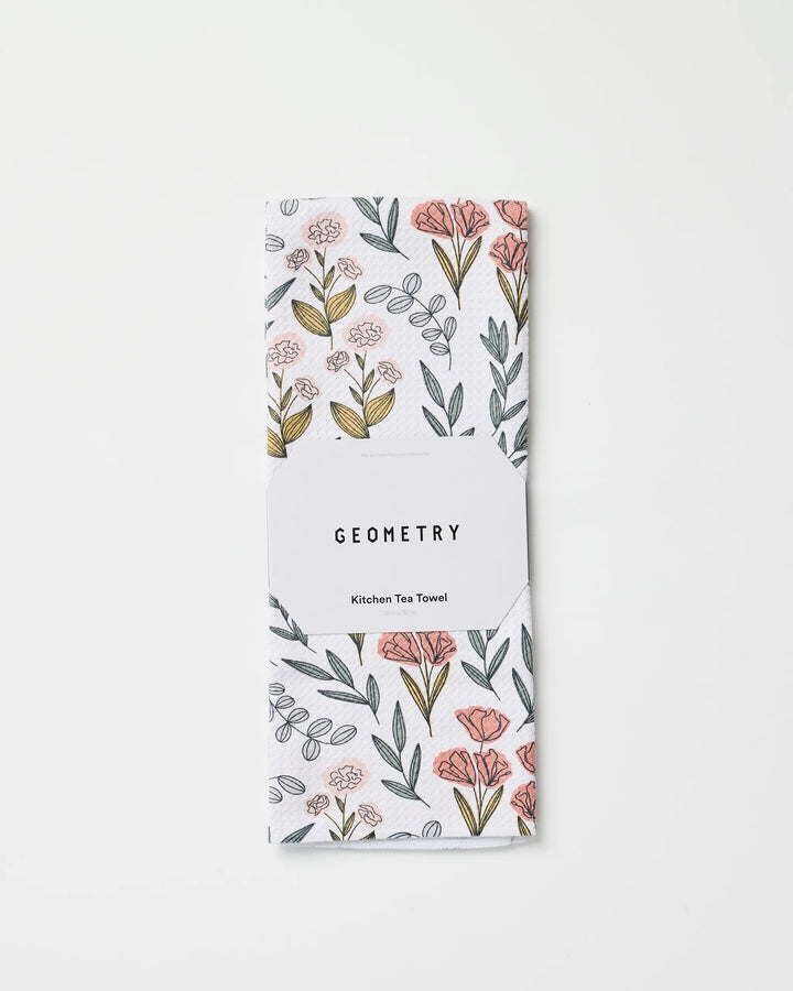 The Charolette Multi tea towel by Charlie Rowan Designs features ultra-absorbent fabric with a pink and yellow floral pattern, folded and wrapped in a white GEOMETRY Kitchen Tea Towel label, displayed on a plain white background.