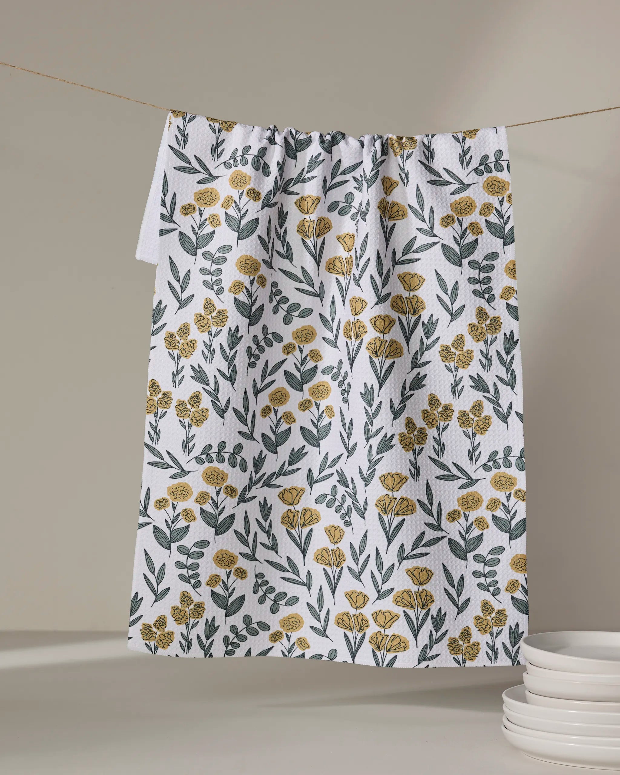 The Charolette Yellow tea towel by Charlie Rowan Designs, featuring a yellow and green floral pattern, hangs on a clothesline near a stack of white plates against a neutral background.