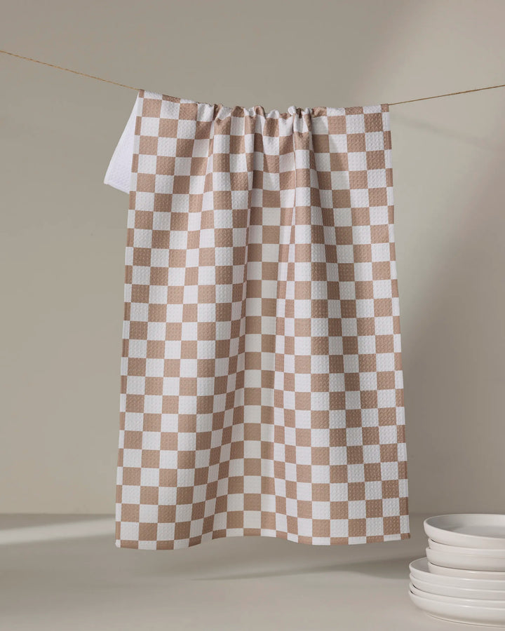 Kelsi Fullmer's Check Mate, a beige and white checkered tea towel, hangs on a string against a neutral background with a stack of white plates in the lower right corner.
