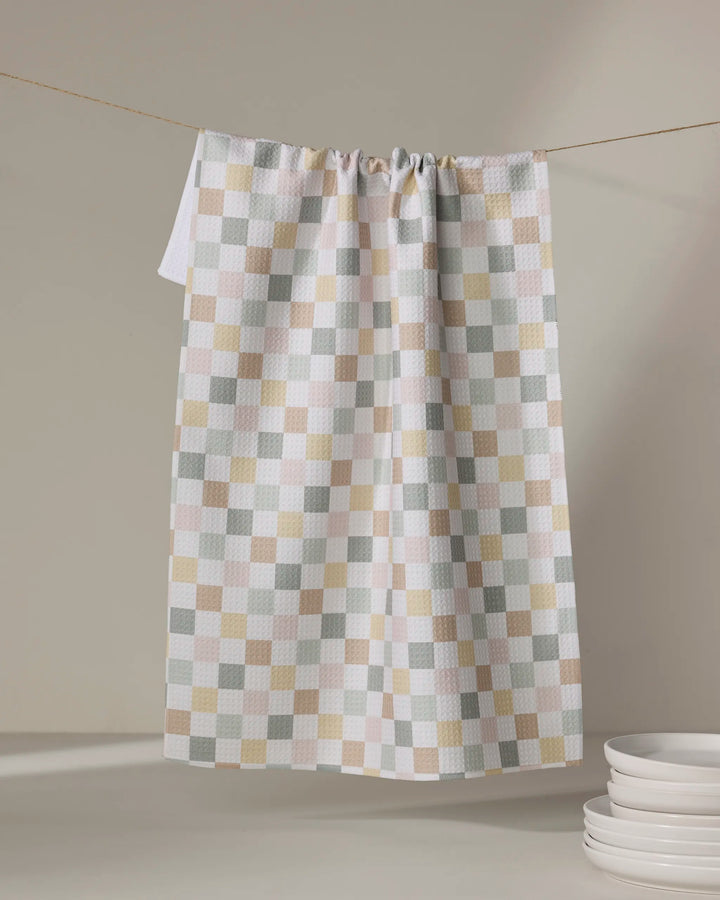 The "Check Me Out" tea towel by Kelsi Fullmer, featuring a beige, gray, white, and light green checkered design, hangs on a string. Made from ultra-absorbent recycled materials. Nearby are stacked white plates.