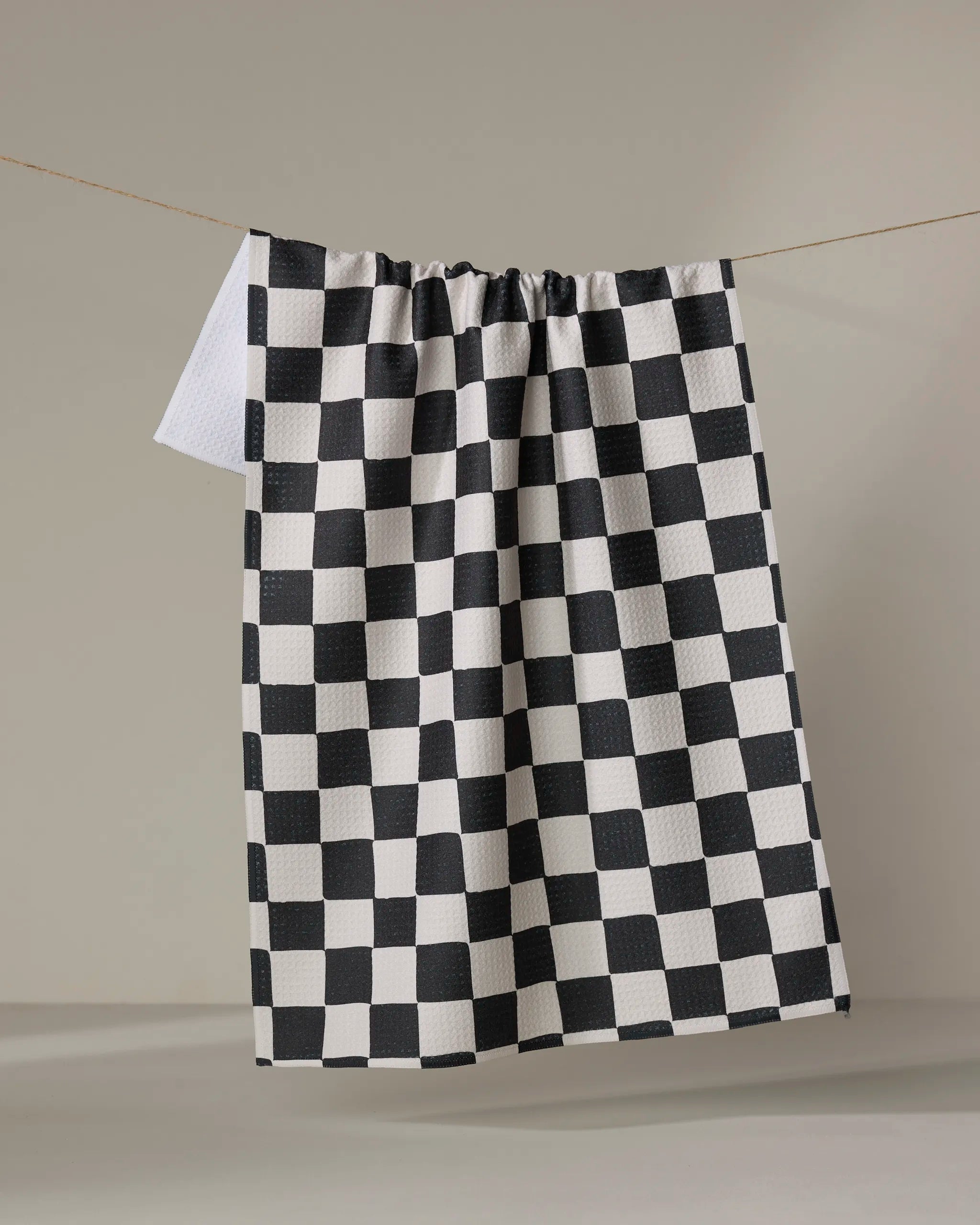 The Checkmate kitchen towel by Danelys Sidron features a classic black and white checkerboard pattern and hangs against a light background. Crafted from recycled materials, it combines style and sustainability.