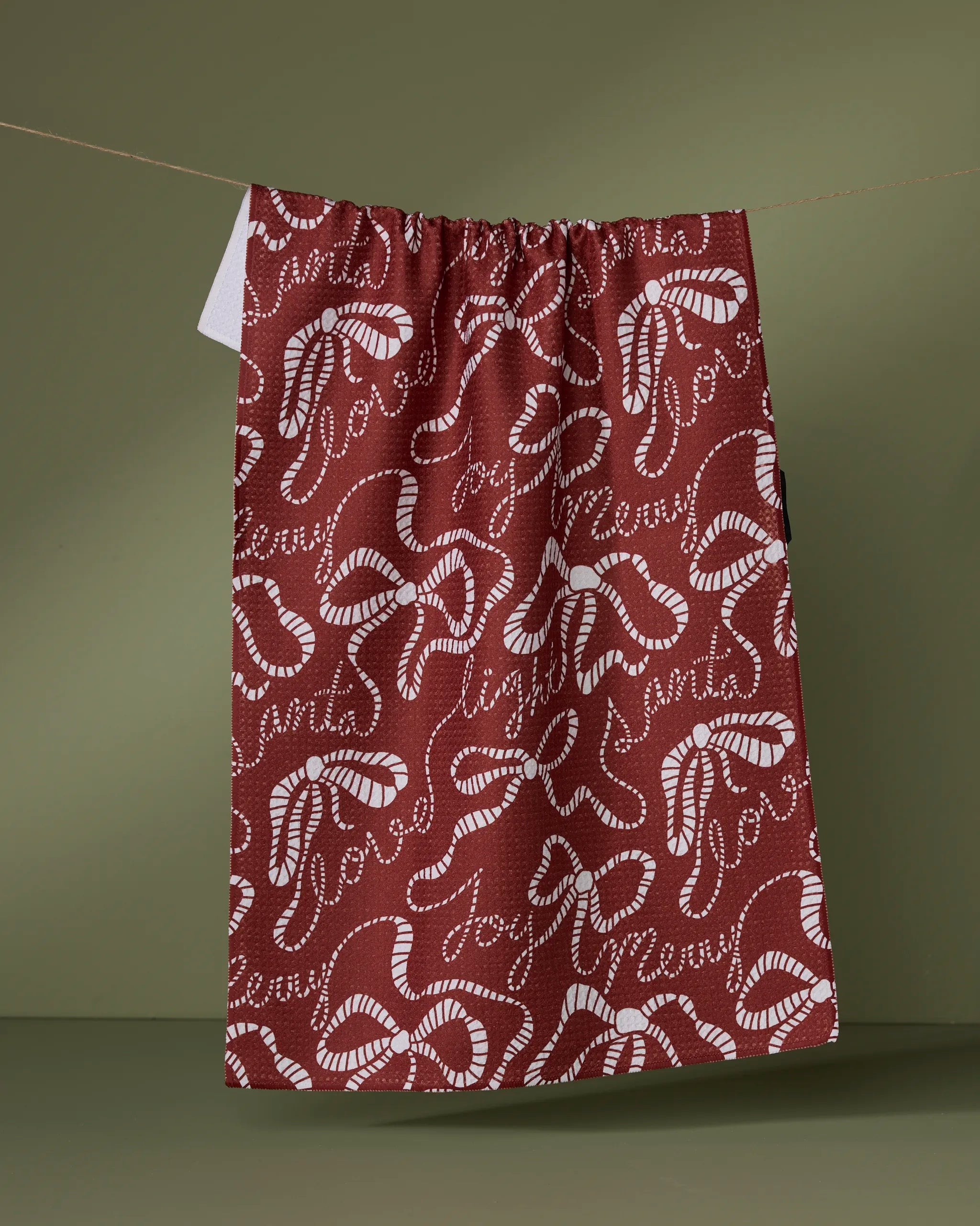 The Geometry "Cheerful Bows" kitchen towel, made from recycled materials, features a maroon color with a white abstract squiggle pattern and hangs by a string against an olive green background.