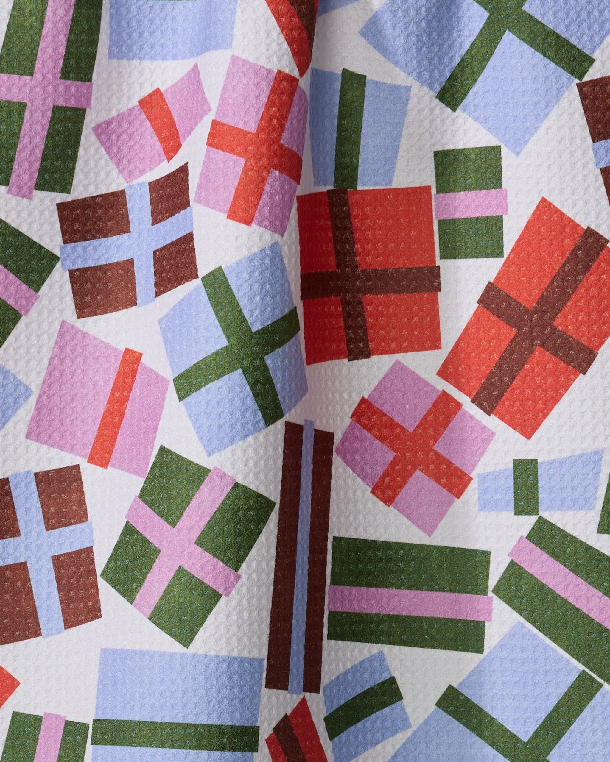 A close-up of the Cheerful Presents recycled kitchen towel by Claire Iglesias, featuring a colorful geometric pattern of gift-like rectangles and squares in red, purple, green, and blue on a white textured background.
