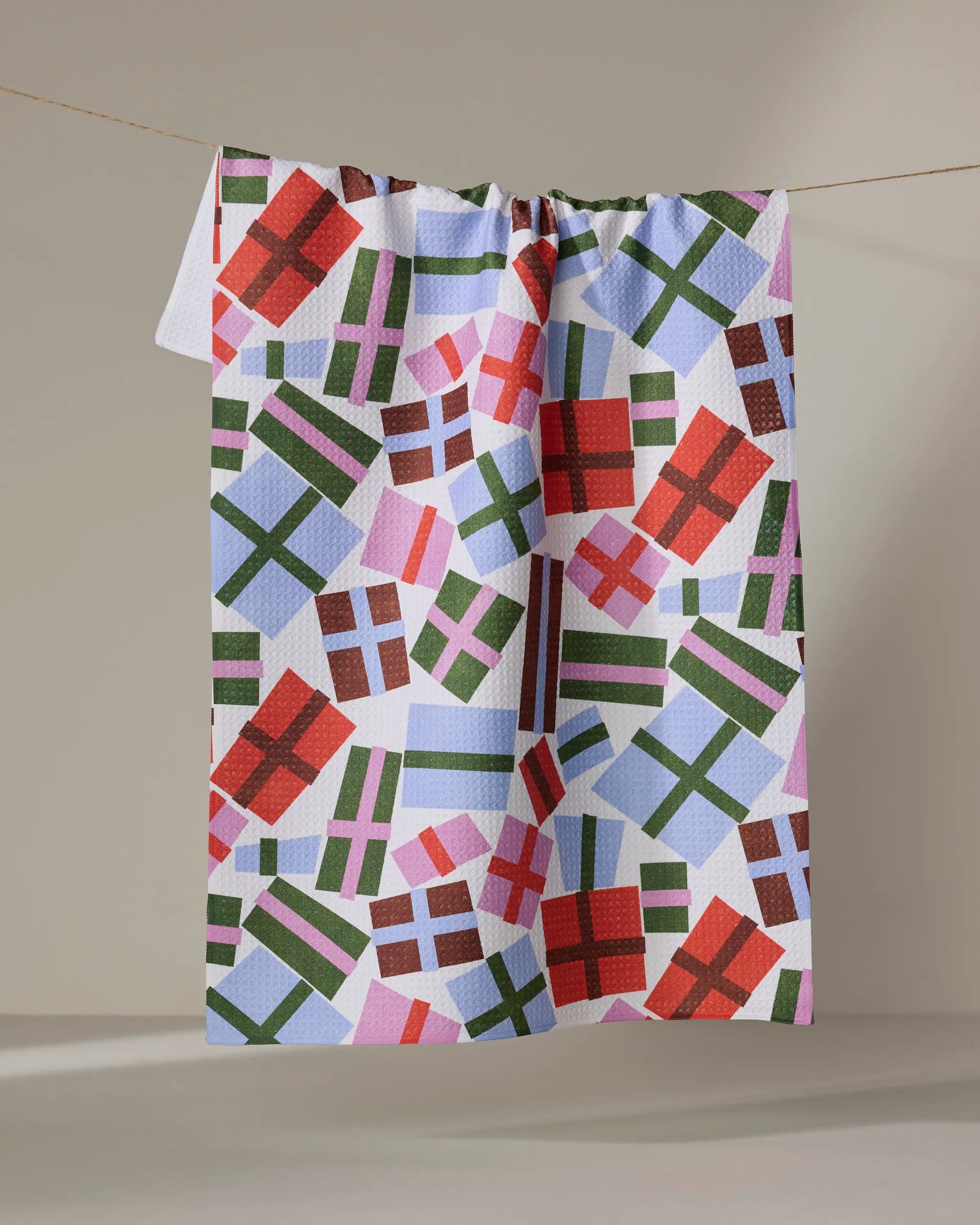 The Claire Iglesias "Cheerful Presents" recycled kitchen towel, featuring vibrant red, green, and purple gift box patterns, hangs from a clothesline against a simple light background.