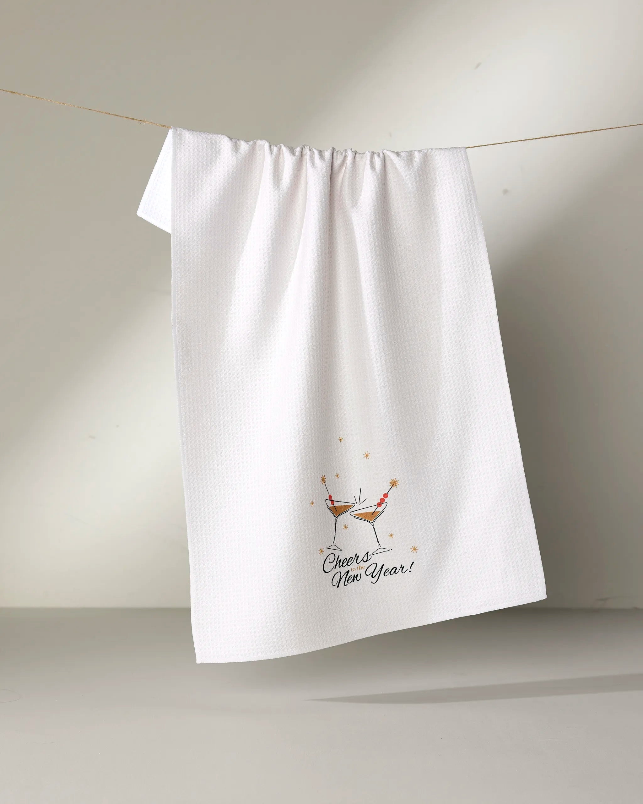 A white textured kitchen tea towel from Geometry, called "Cheers," hangs on a line. Made from recycled materials, it features a festive drink with bubbles, orange stars, and “Cheers! New Year!” near the bottom.