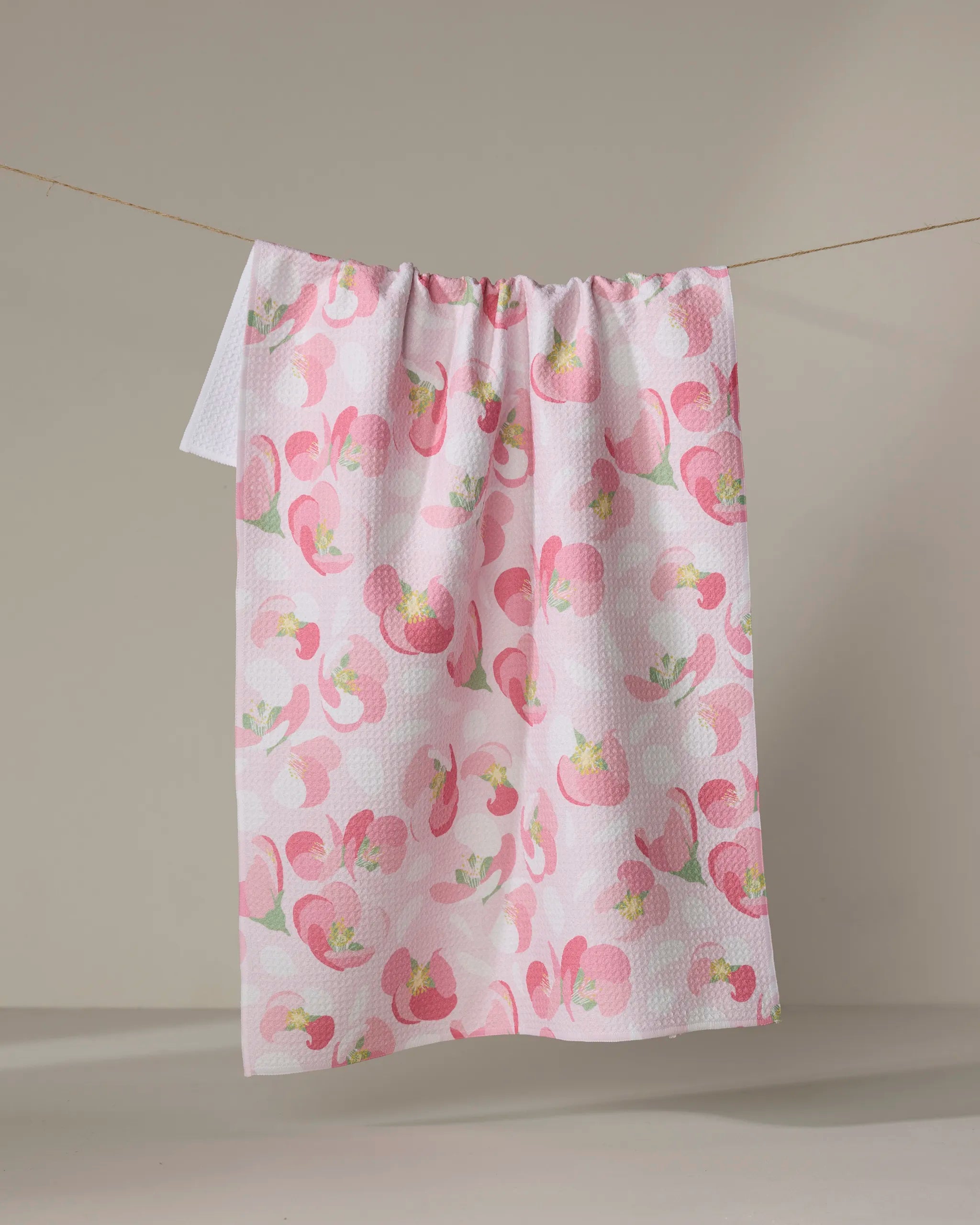 The Rebecca Bobko "Cherry Blossom" white tea towel, featuring a pink and green floral pattern and made from recycled materials, hangs on a line against a plain beige background.