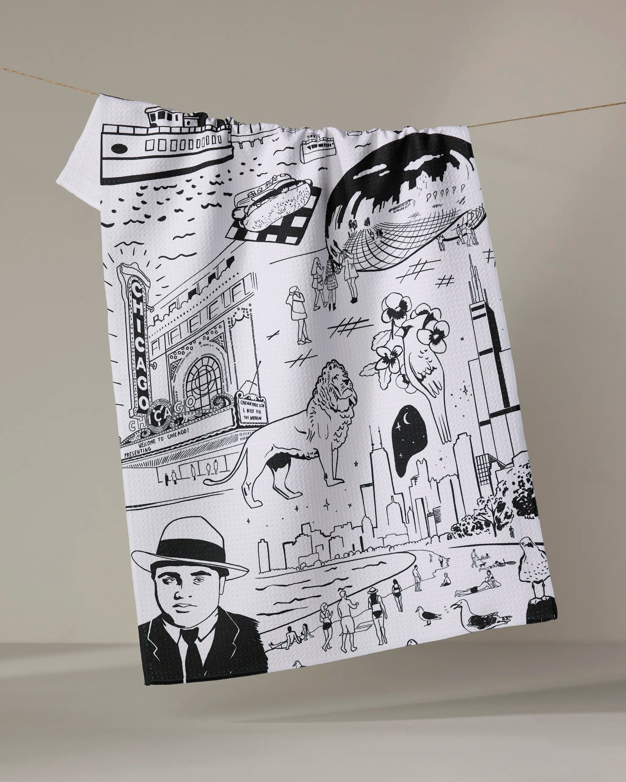 The Elisabeth Wing "Chicago" tea towel features black-and-white illustrations of the skyline, a lion, Chicago Theater, boat, clouds, and people. Made from recycled materials and shown hanging on a line against a neutral background.