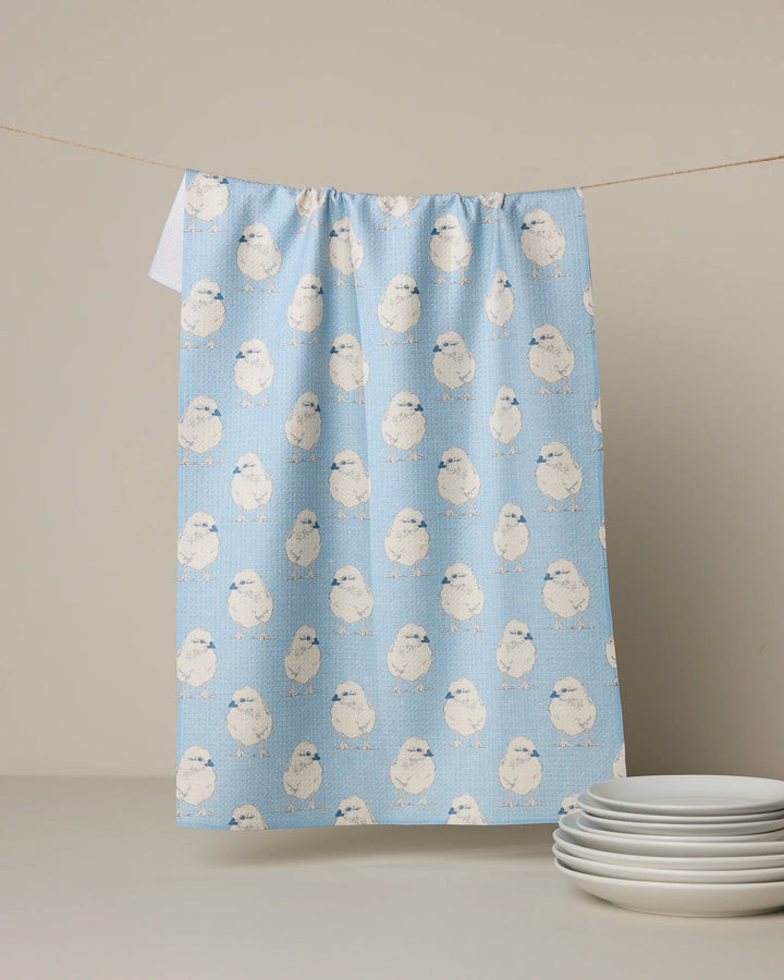 A Geometry Chickadee recycled kitchen towel, featuring a blue design with a white bird pattern, hangs on a line above stacked white plates on a white surface.