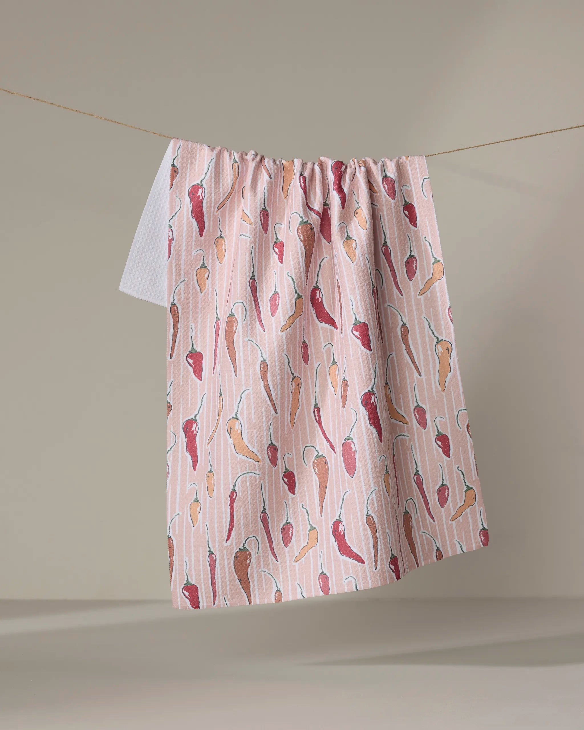 The Chilli Stripe tea towel by Hufton Studio is ultra-absorbent and pink, featuring illustrated red, orange, and brown chili peppers. It hangs on a clothesline against a neutral background.