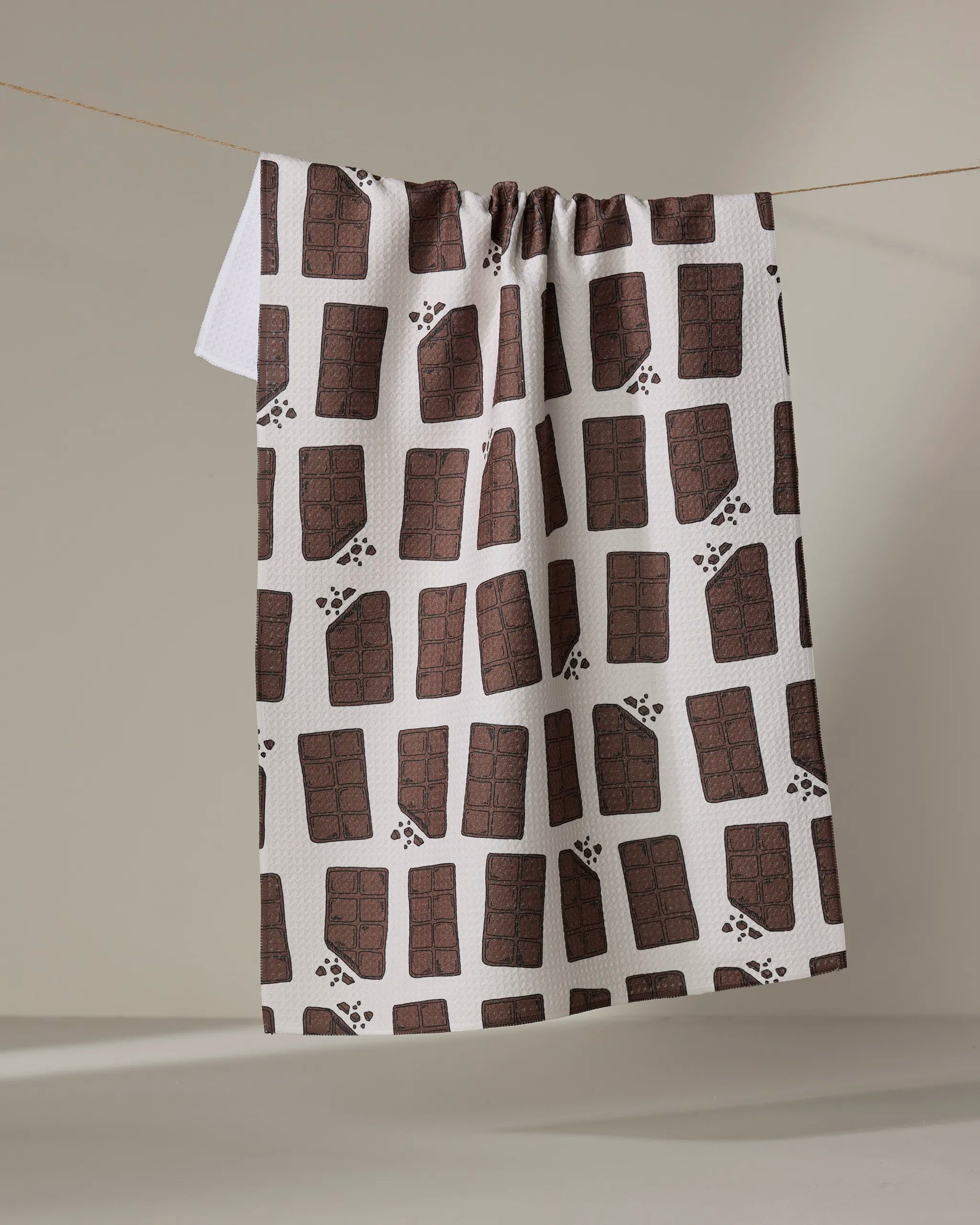 The "Chocolate Everything" tea towel by Amy Droubay, made from recycled materials, displays a brown chocolate bar pattern with brown paw prints and hangs on a clothesline against a neutral background.