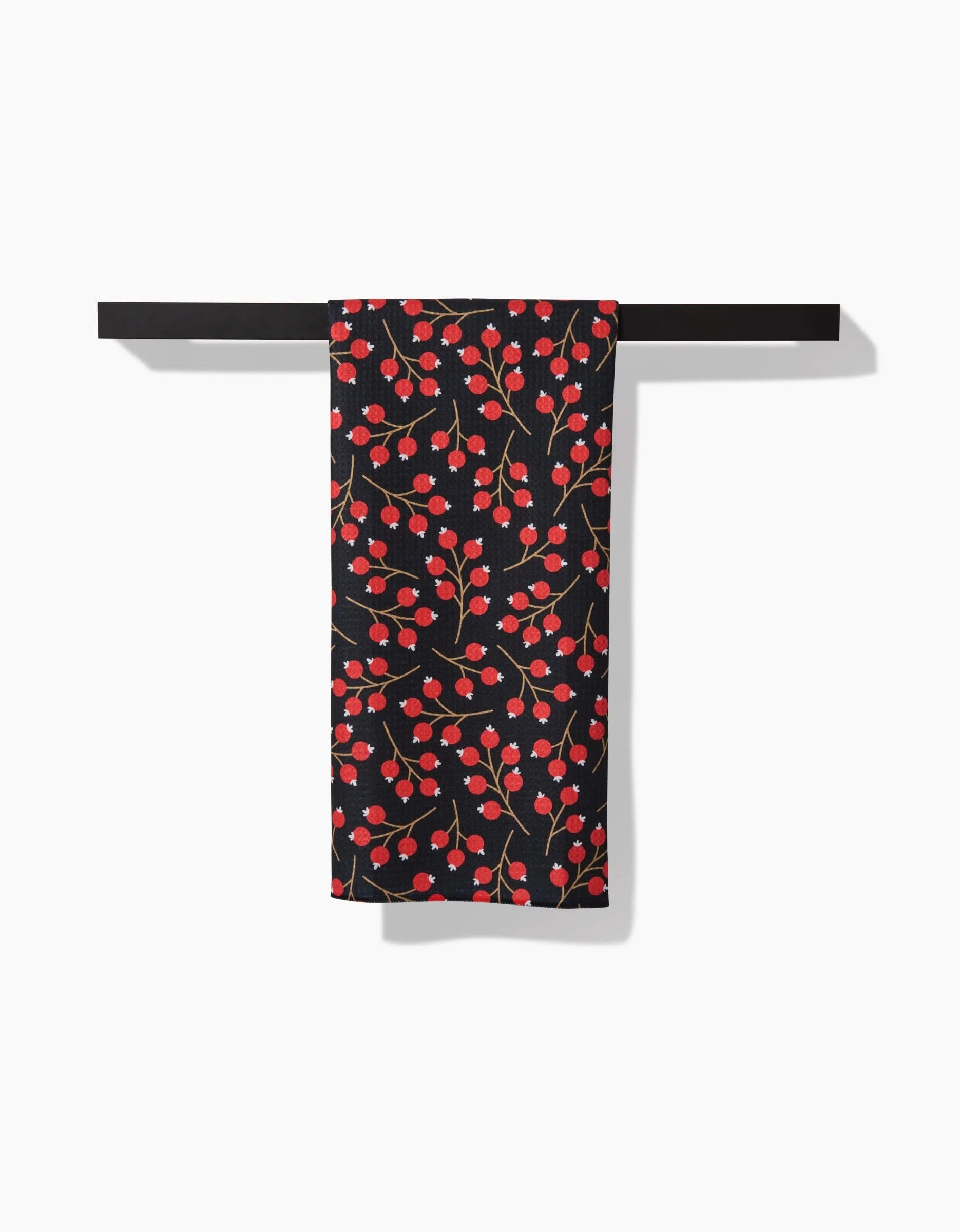 The Megan Kampa Christmas Berries dish towel, featuring a black absorbent fabric with a red and pink floral pattern, hangs neatly over a horizontal black bar against a white background.