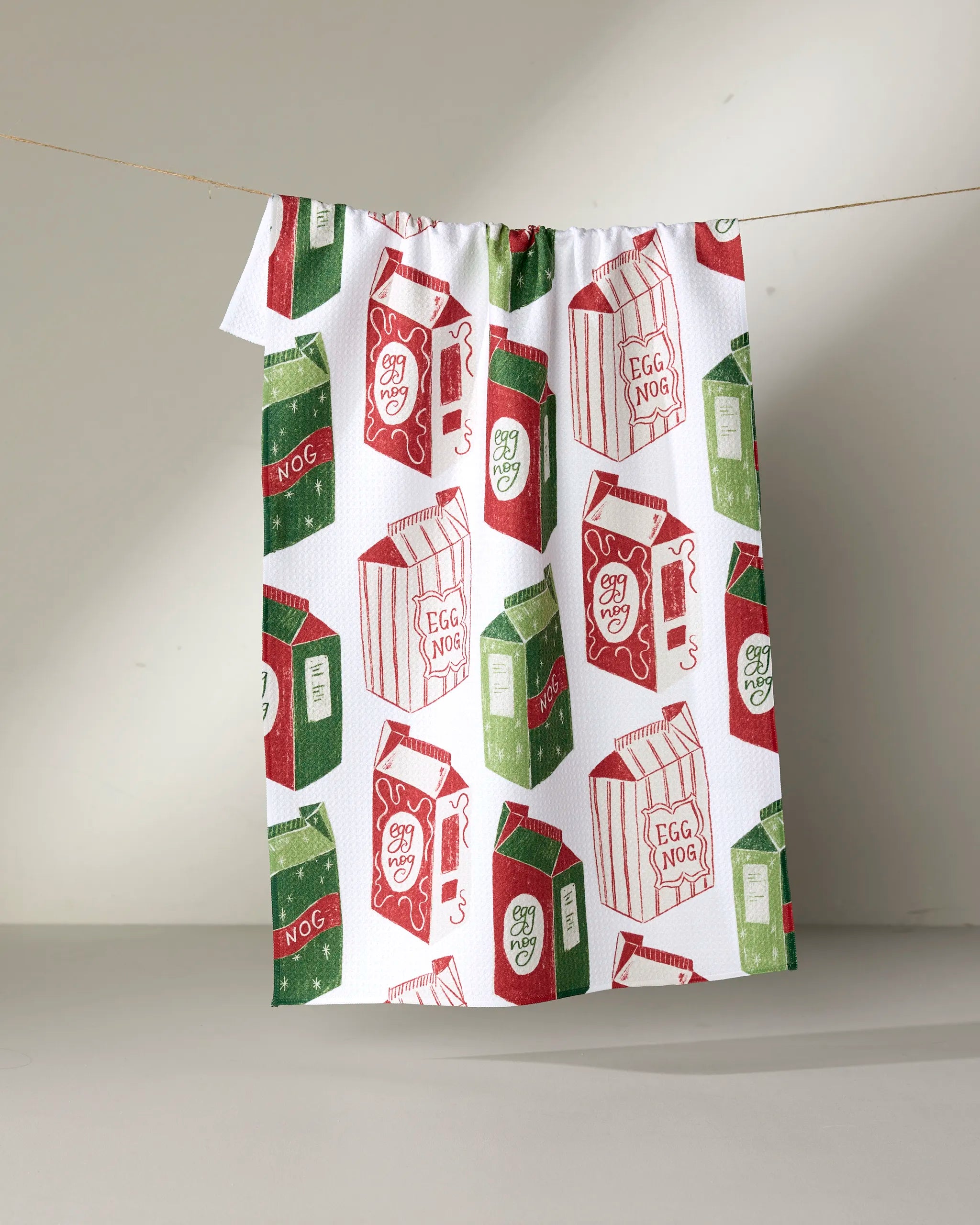 The Aubrey Fairchild "Christmas Eggnog" recycled kitchen towel features red and green eggnog cartons hanging on a clothesline against a neutral background.