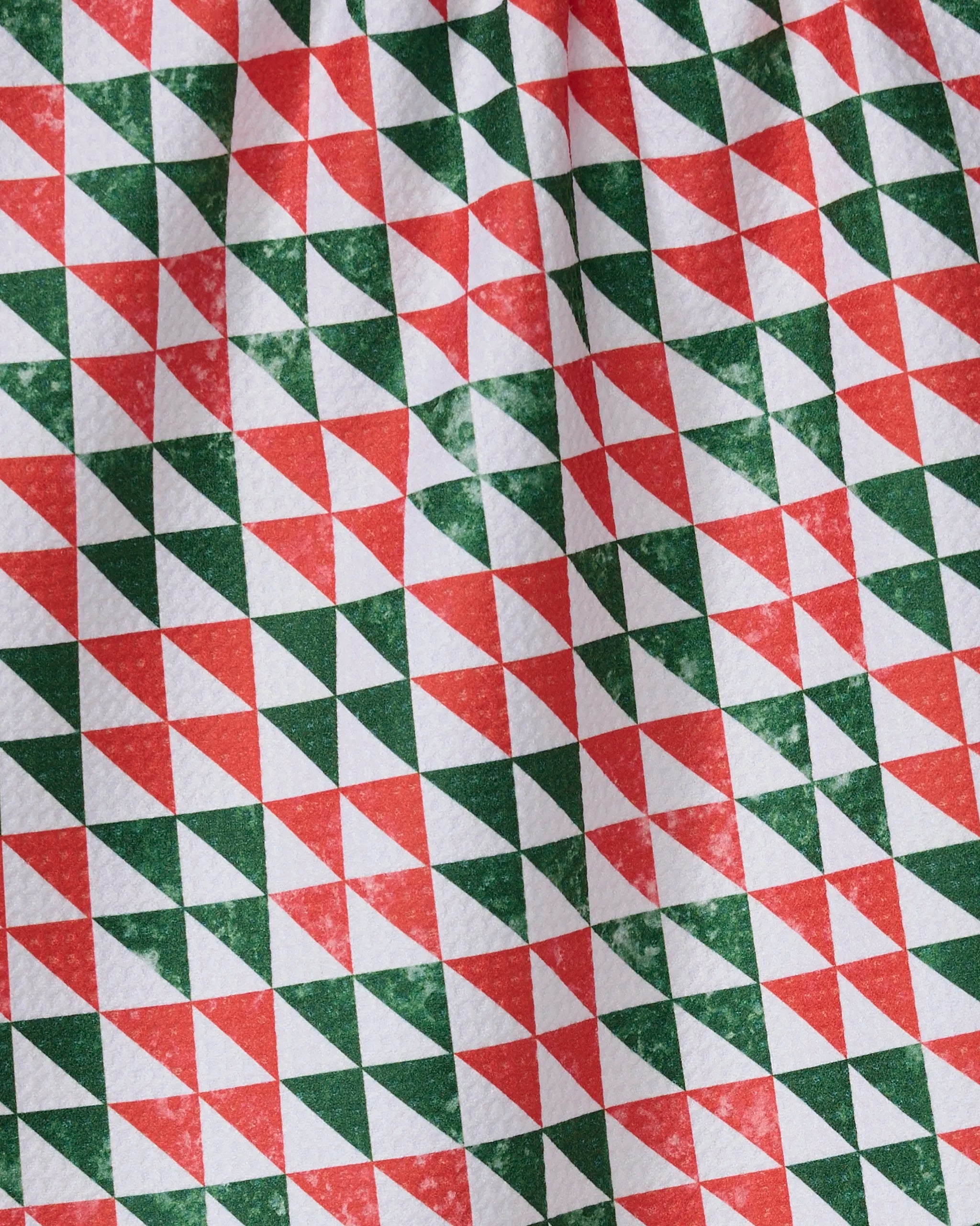 The Geometry Christmas Evergreen kitchen towel showcases a geometric pattern of red and green triangles forming diamonds on a white background. Made from recycled materials, its fabric has a slightly wrinkled appearance.