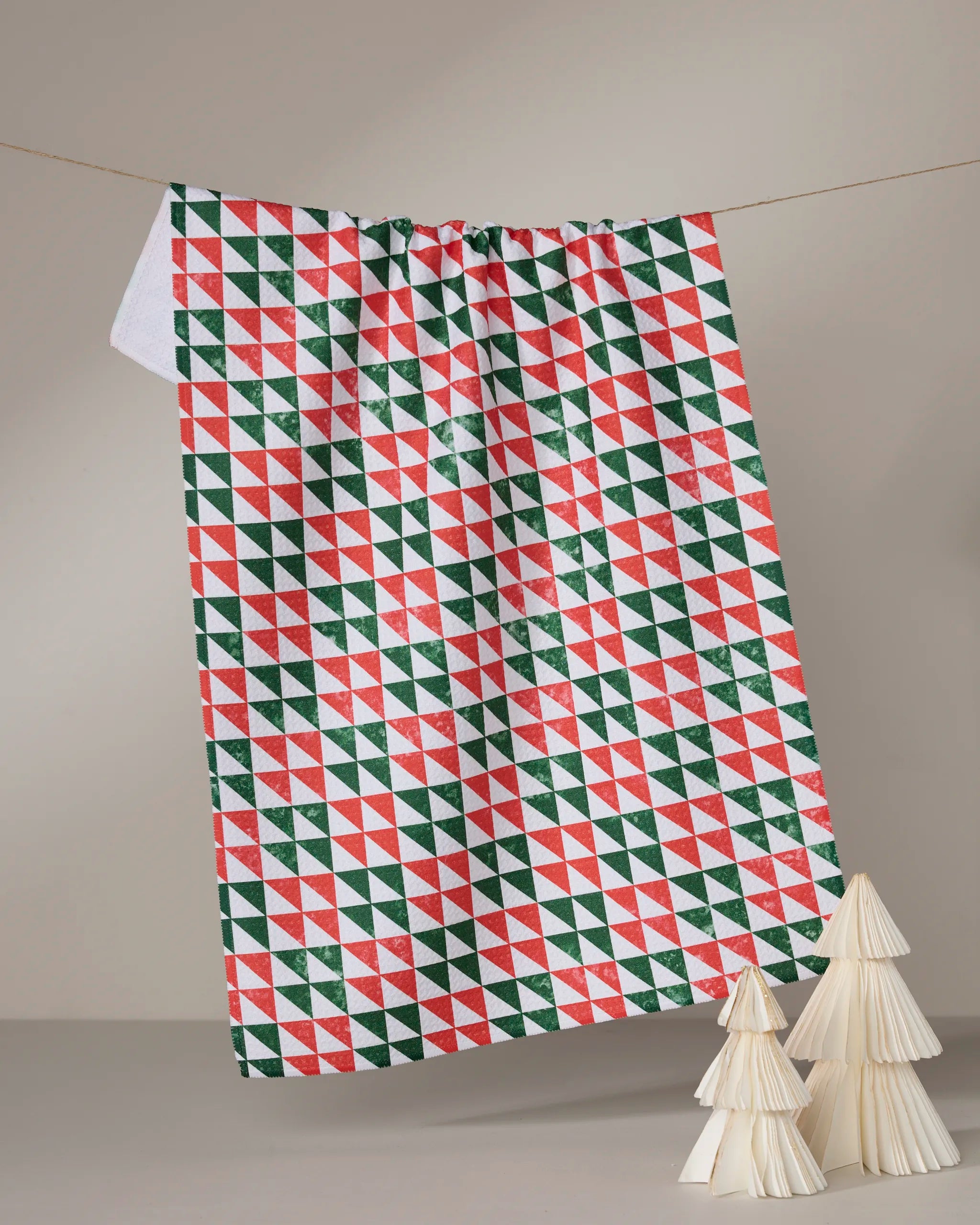 The Geometry Christmas Evergreen tea towel, featuring a red, green, and white geometric triangle pattern, hangs on a line with two small white decorative cone trees in the foreground.