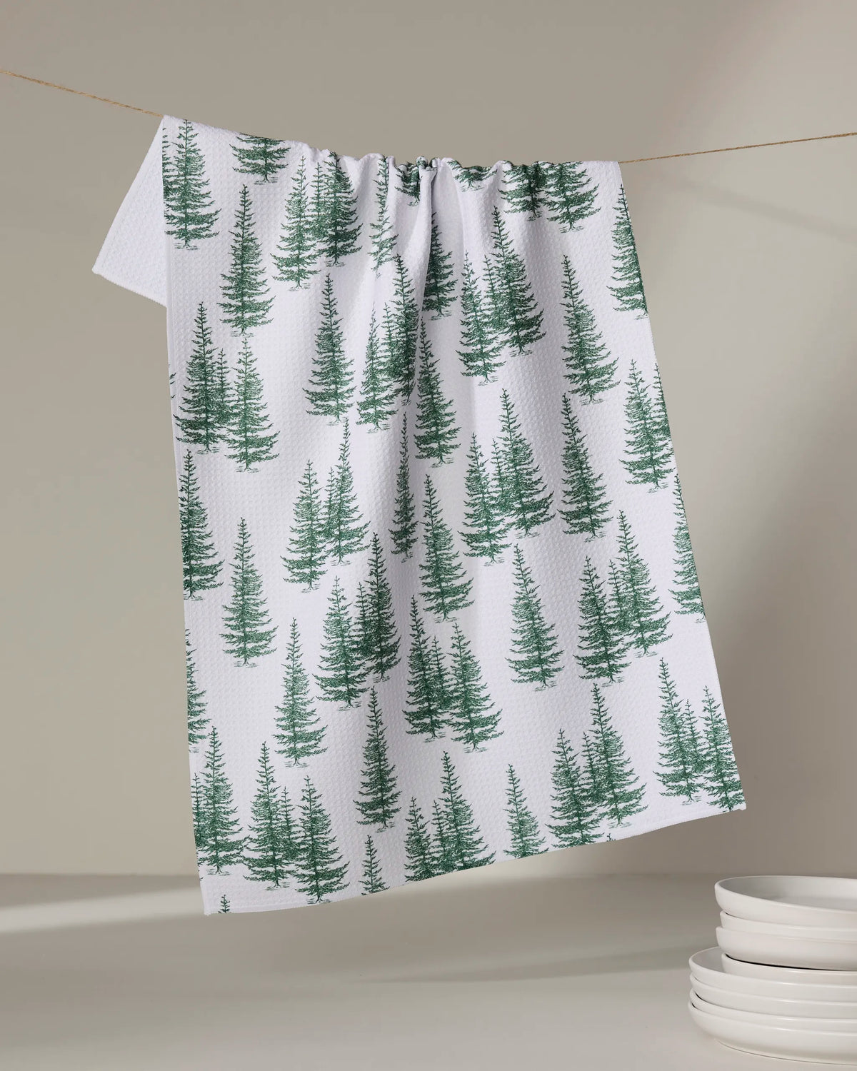 The Aubrey Fairchild "Christmas Forest" tea towel, made from recycled materials and featuring green pine trees, hangs on a clothesline with white plates stacked in the softly lit background, adding eco-friendly charm to your kitchen.