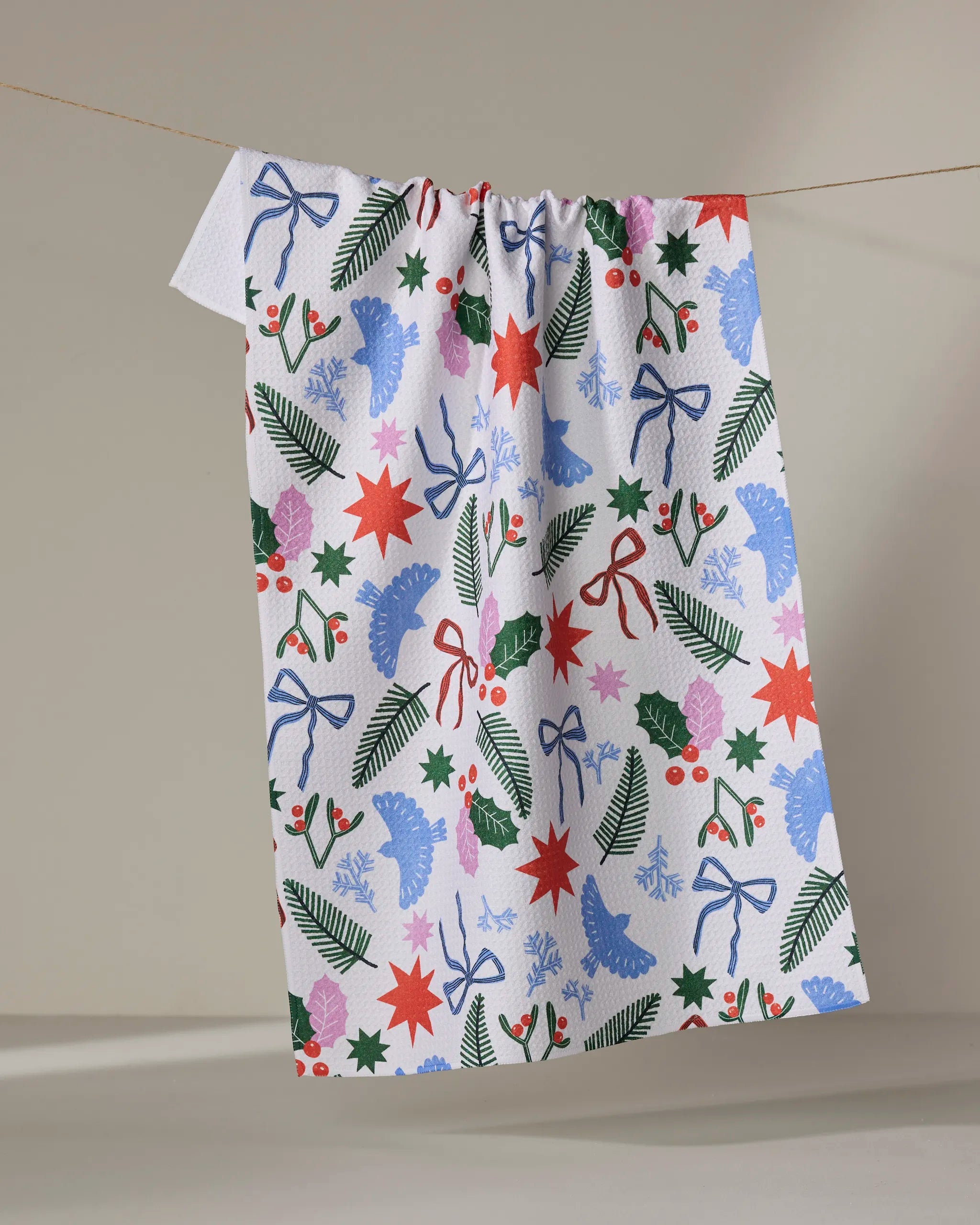 The Claire Iglesias “Christmas Garden Party” dish towel features a festive holiday pattern—blue bells, red stars, holly, berries, pine, and blue ribbons—displayed on a clothesline against a neutral background.