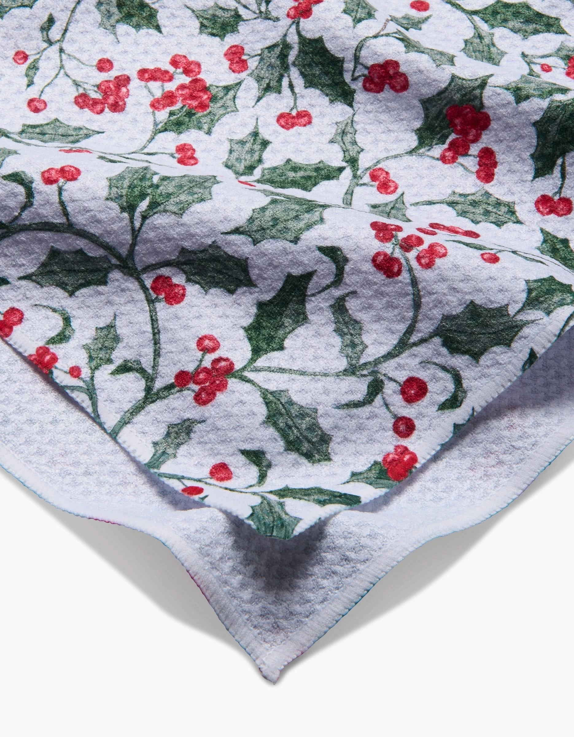 Aubrey Fairchild’s Christmas Holly tea towel features a white textured fabric with green holly leaves and red berry clusters, shown partially folded on a white background.