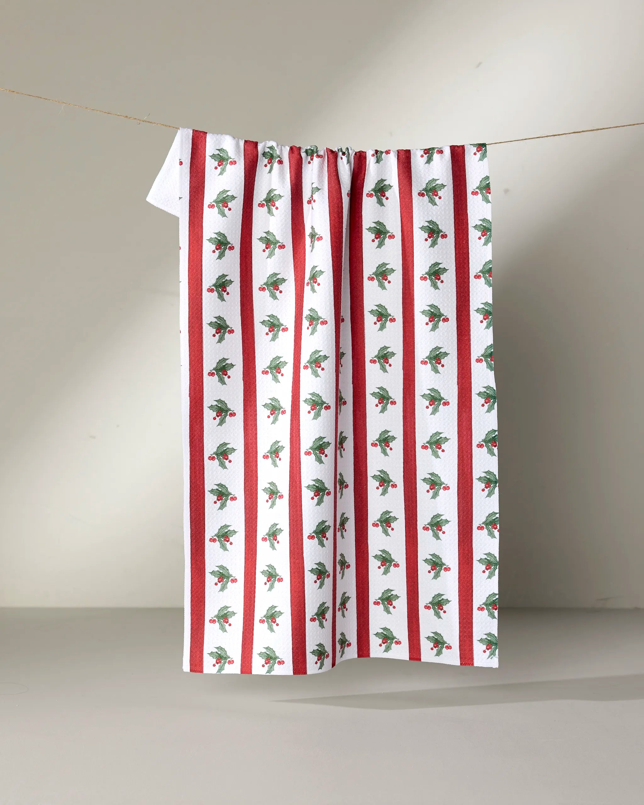 The Aubrey Fairchild Christmas Holly Stripe kitchen towel, featuring eco-friendly white fabric with red vertical stripes and holly patterns, hangs on a line against a neutral background.