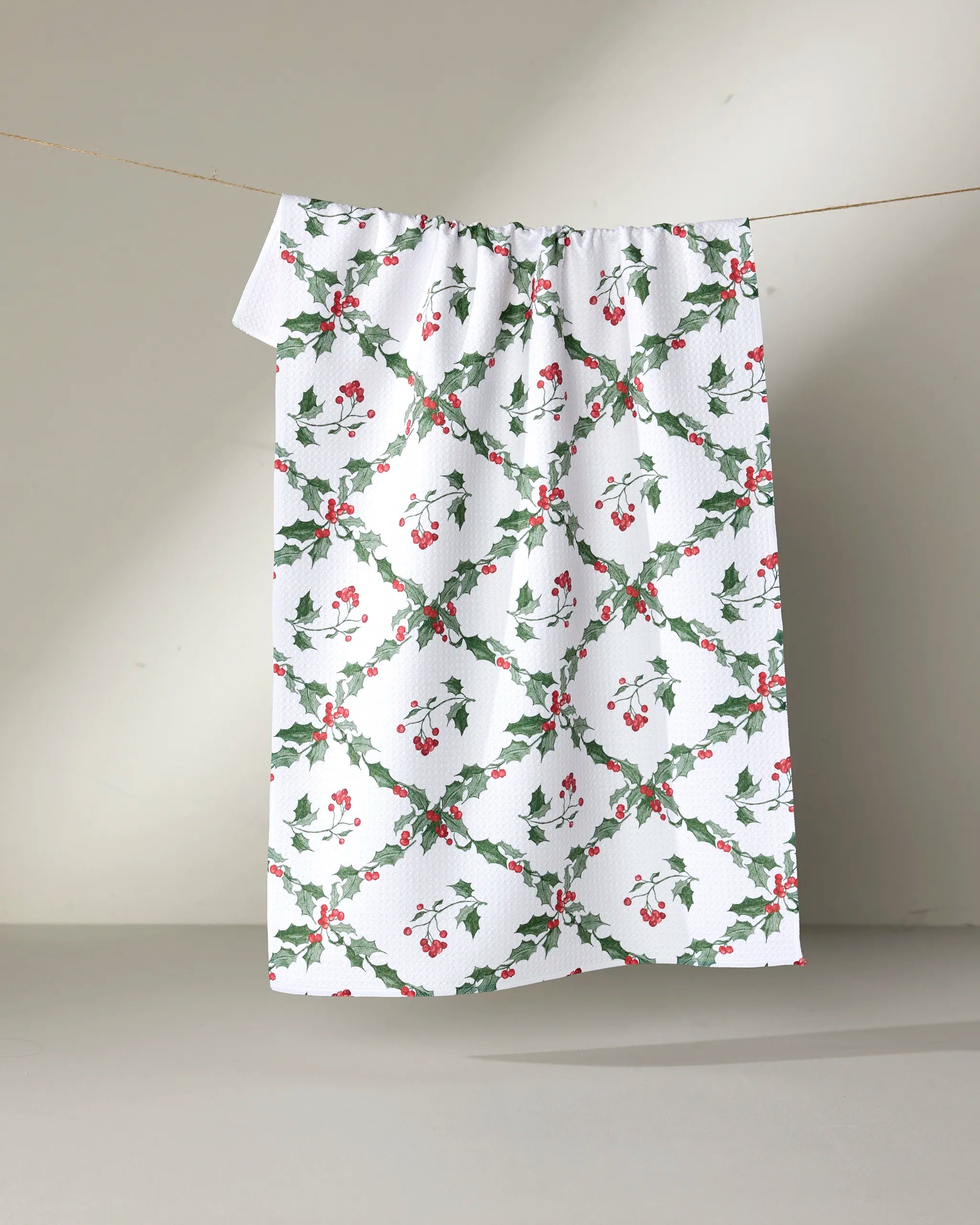 The Aubrey Fairchild "Christmas Lattice" tea towel features green holly leaves and red berries on a neutral background. Festive and absorbent, this holiday-themed towel is made from recycled materials.