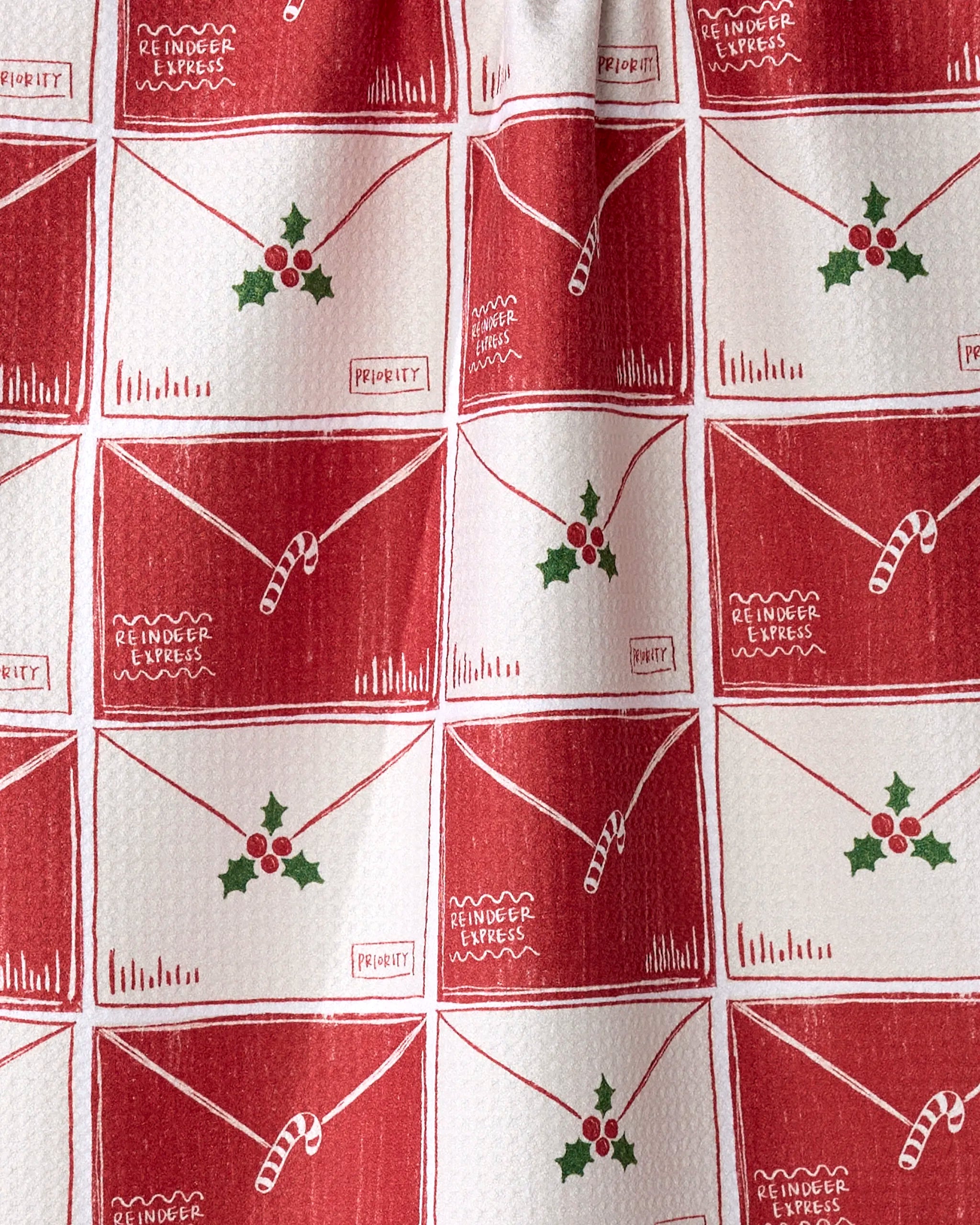 The Christmas Letter Stack tea towel by Aubrey Fairchild features red and white patterned Geoweave fabric with hand-drawn envelopes, holly, candy canes, and “Reindeer Express” details—festive style made from recycled materials.