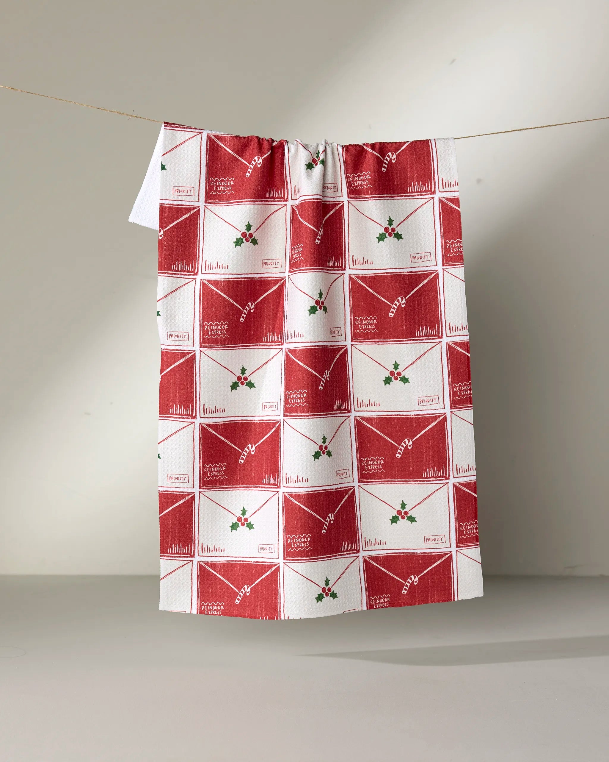The Aubrey Fairchild Christmas Letter Stack tea towel, made from recycled materials, hangs on a line. Its red and white design features envelopes with holly and festive greetings like "Merry Christmas" and "With Love.