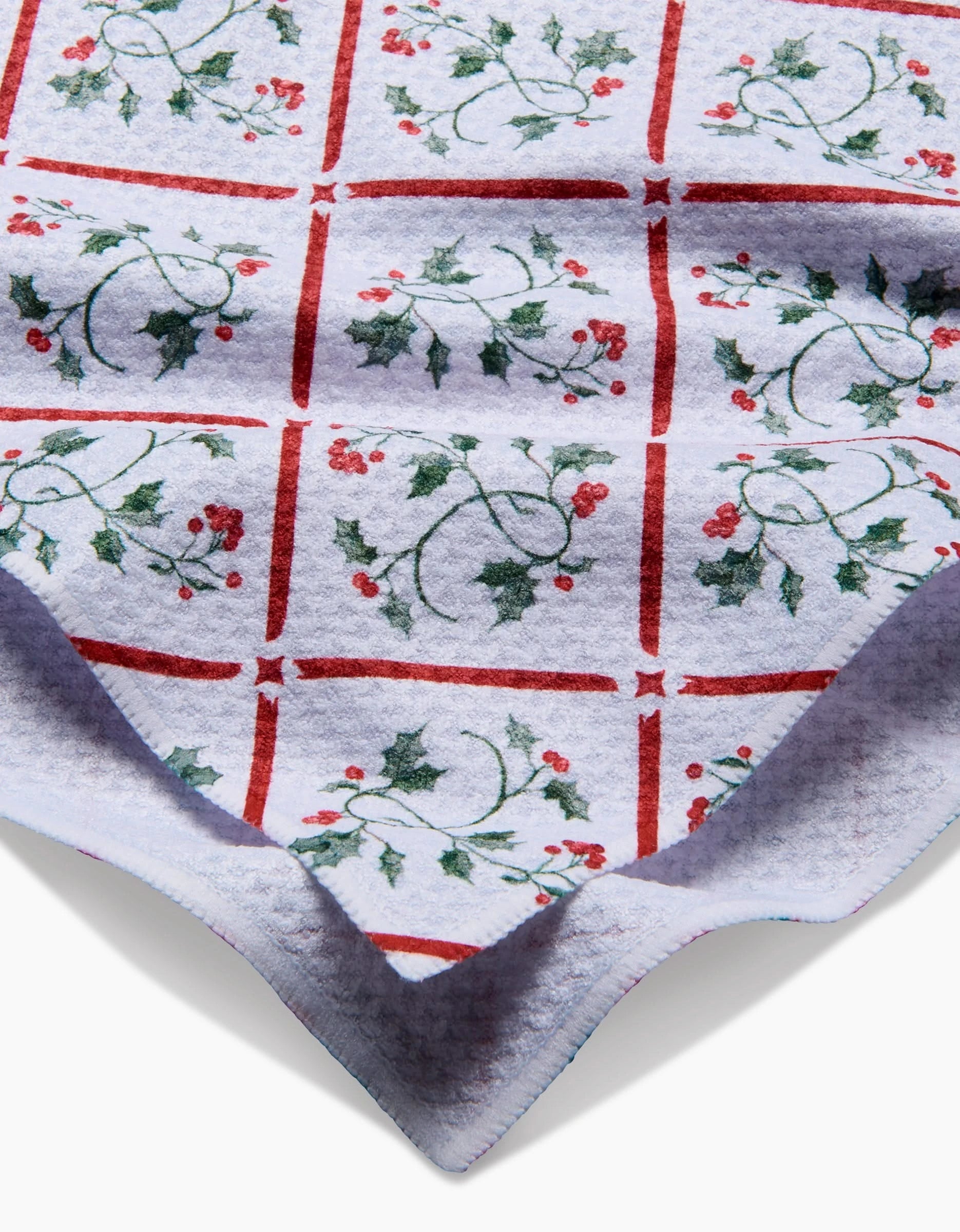 The Aubrey Fairchild Christmas Mistletoe Trellis is a soft white blanket with a red grid, green holly leaves, and red berries in each square. Folded at the corner, it’s crafted from post-consumer recycled materials for eco-friendly comfort.