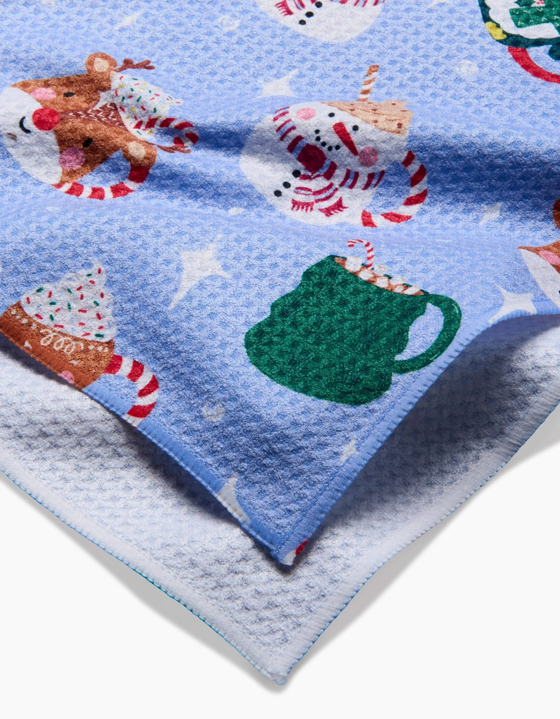 The Christmas Mugs blanket by Vaishali Design Studio features blue textured fabric with festive patterns—reindeer, snowmen, green mugs with candy canes, and holiday treats—recycled materials, a white underside, and a stitched edge.